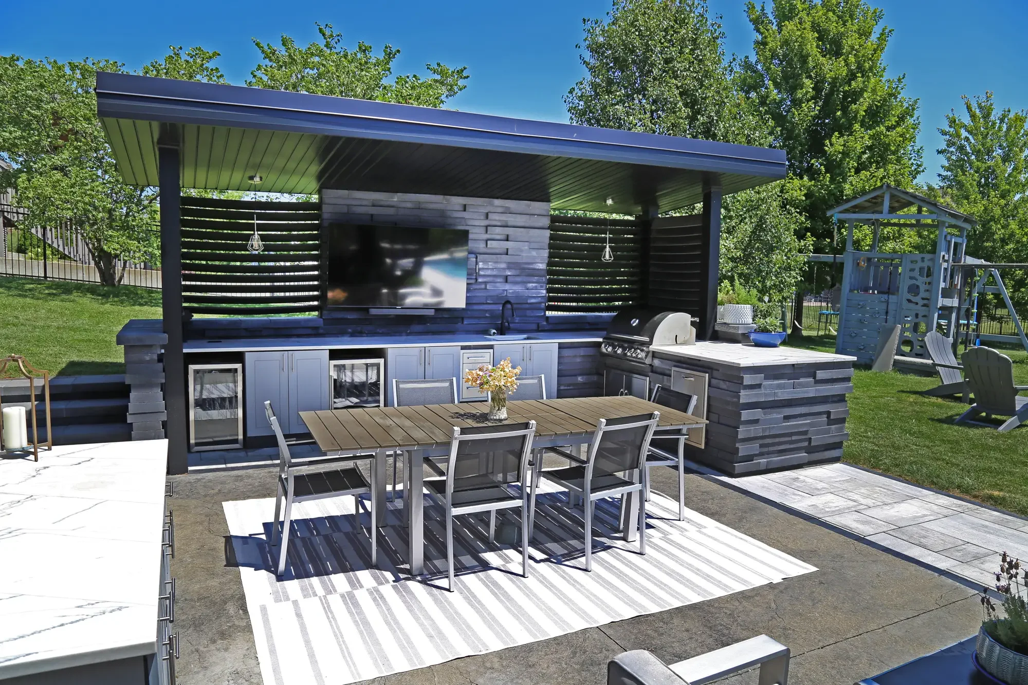 Modern outdoor kitchen with stacked stone surround, outdoor TV, built-in grill, and dining area under dark pergola at Slate and Steel by Green View in Illinois