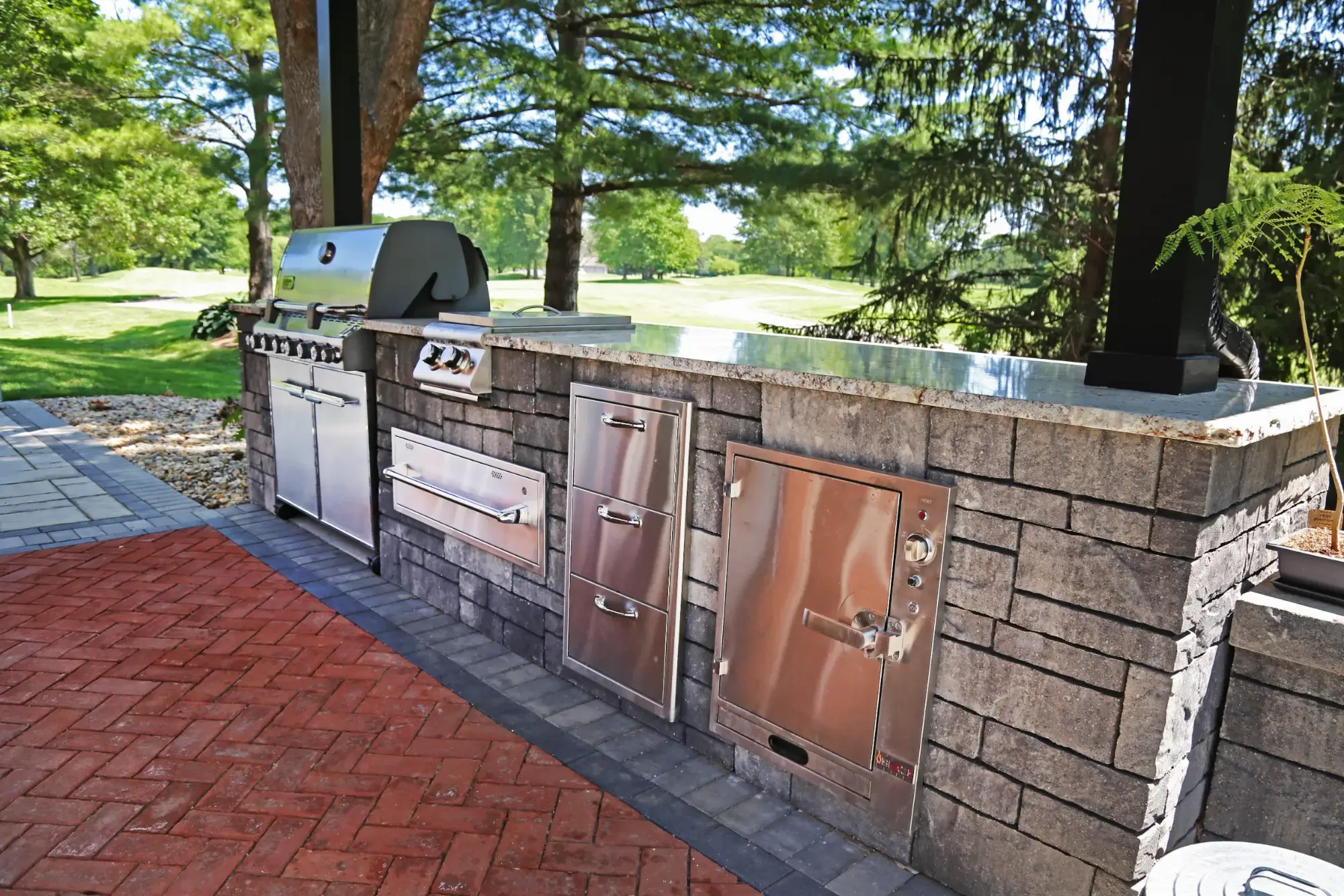 Outdoor kitchen with built-in stainless steel grill and storage cabinets on a stone counter, overlooking a lush green park with trees.
