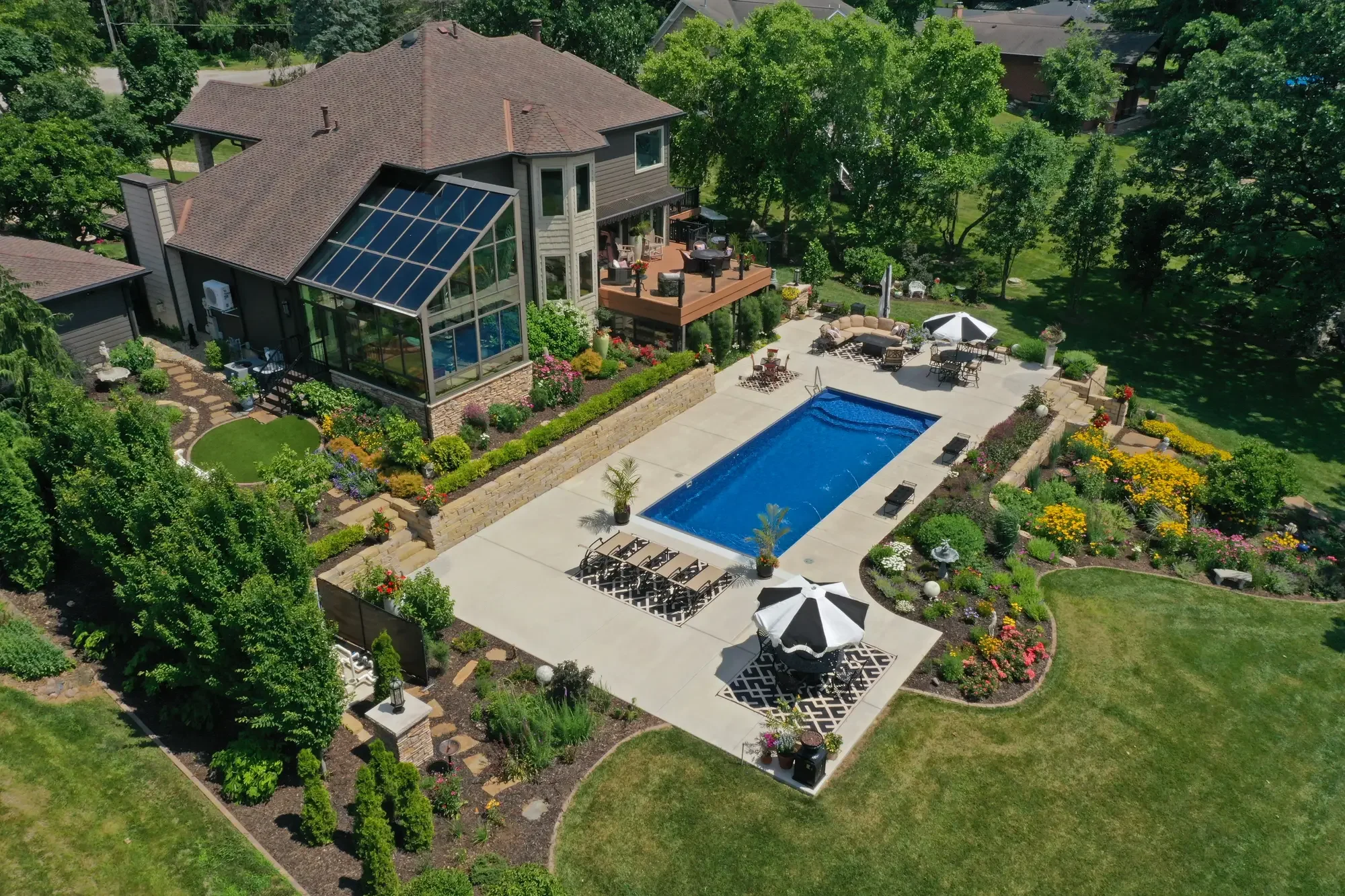 Aerial view of a backyard with a swimming pool, garden, patio furniture, and a house with a sunroom and outdoor deck.