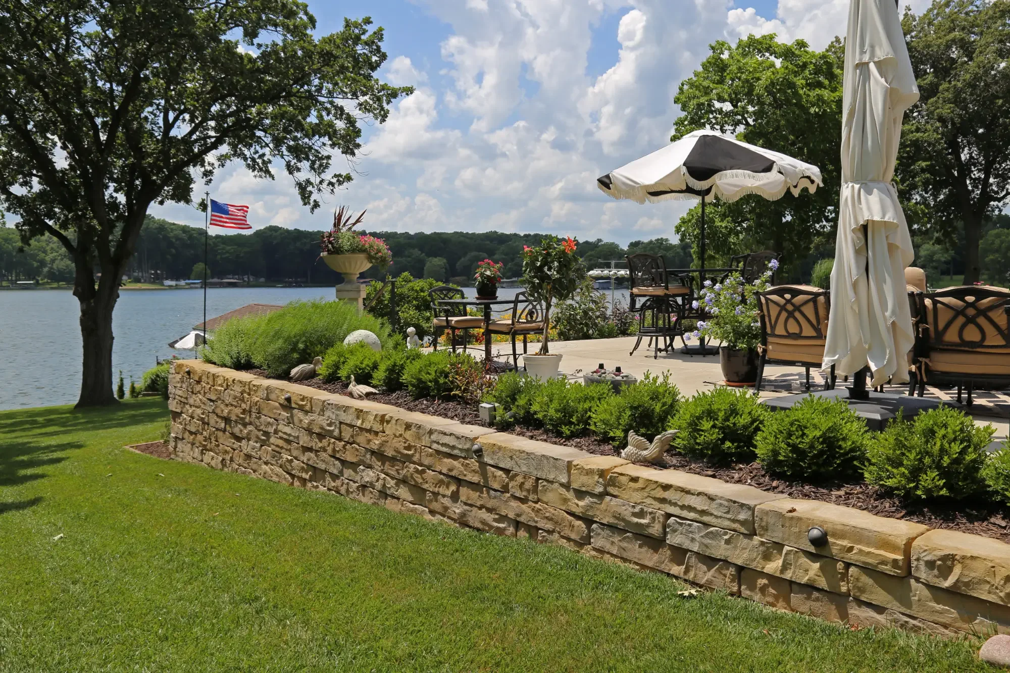 A lakeside patio with outdoor furniture, potted plants, and an American flag, surrounded by green trees and grass under a partly cloudy sky.