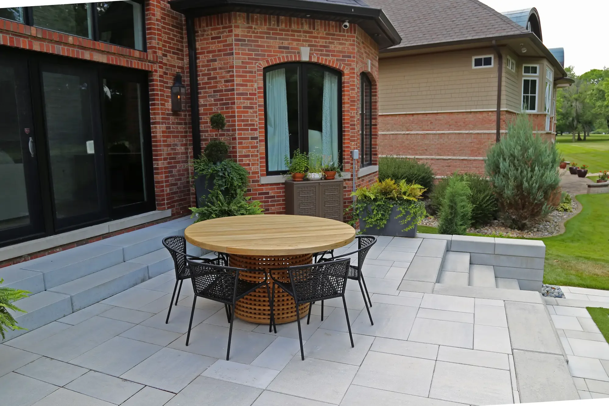 Upper patio dining area with round teak table, metal chairs, and stone seating wall at Geometric Oasis Retreat by Green View in Illinois