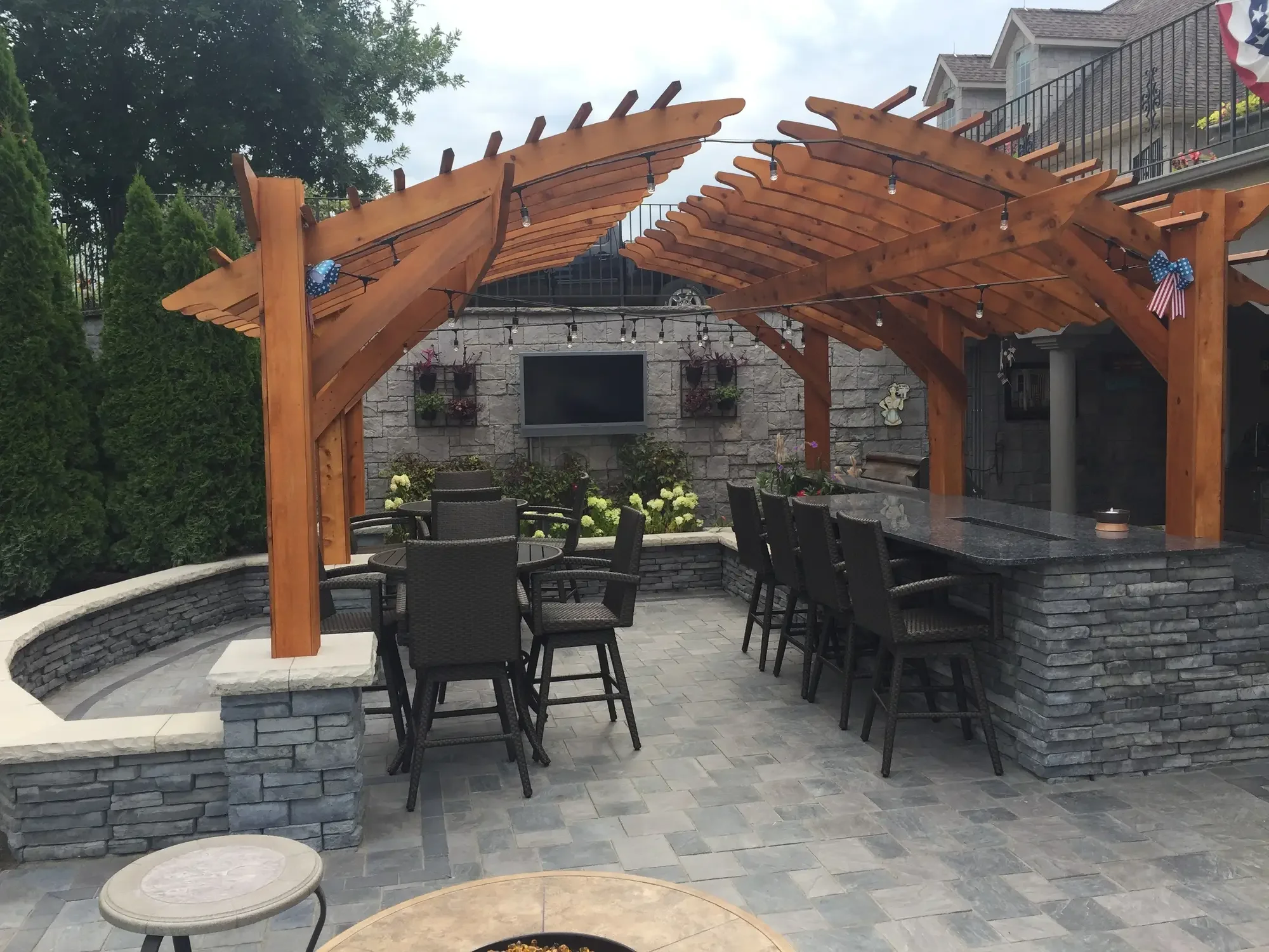 Full view of arched cedar pergola with granite bar, bar dining, curved stone seating wall, and outdoor TV wall at The Craftsman's Retreat by Green View in Illinois