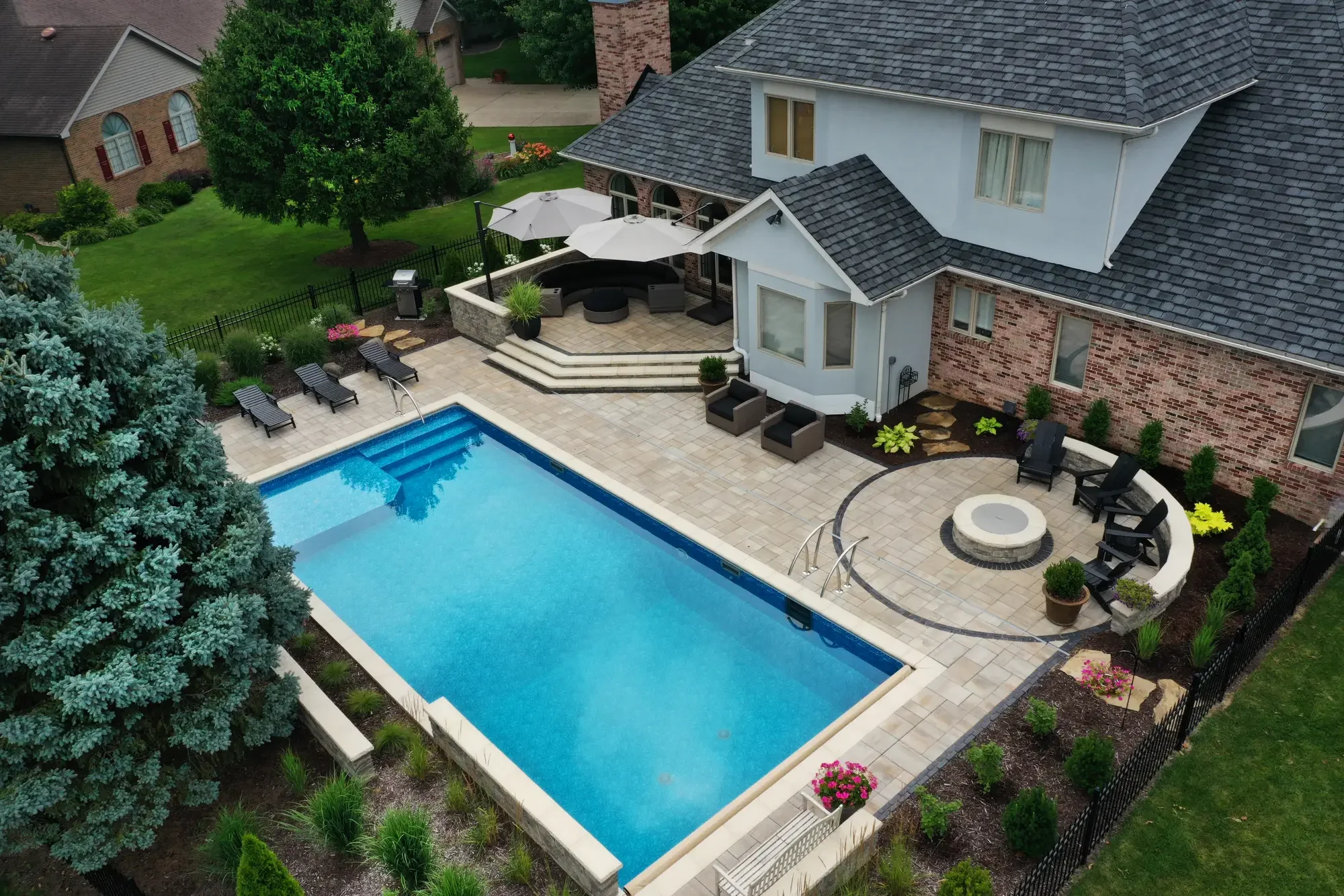 Aerial view of a backyard highlighting a rectangular swimming pool with blue water, surrounded by a beige stone patio. The patio features lounge chairs, potted plants, a fire pit with chairs, and an outdoor seating area with umbrellas. The house has a brick and light-colored exterior with large windows, and is bordered by well-maintained landscaping and greenery.