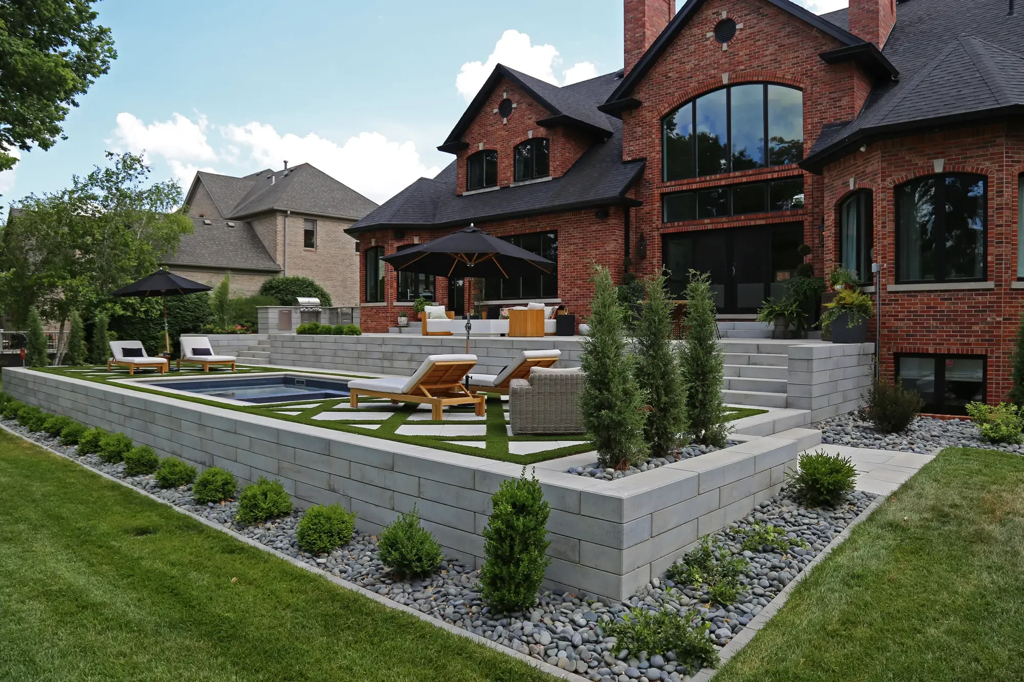 Side yard view of pool terrace with geometric turf-and-paver surround, tiered stone walls, and river rock planting border at Geometric Oasis Retreat by Green View in Illinois