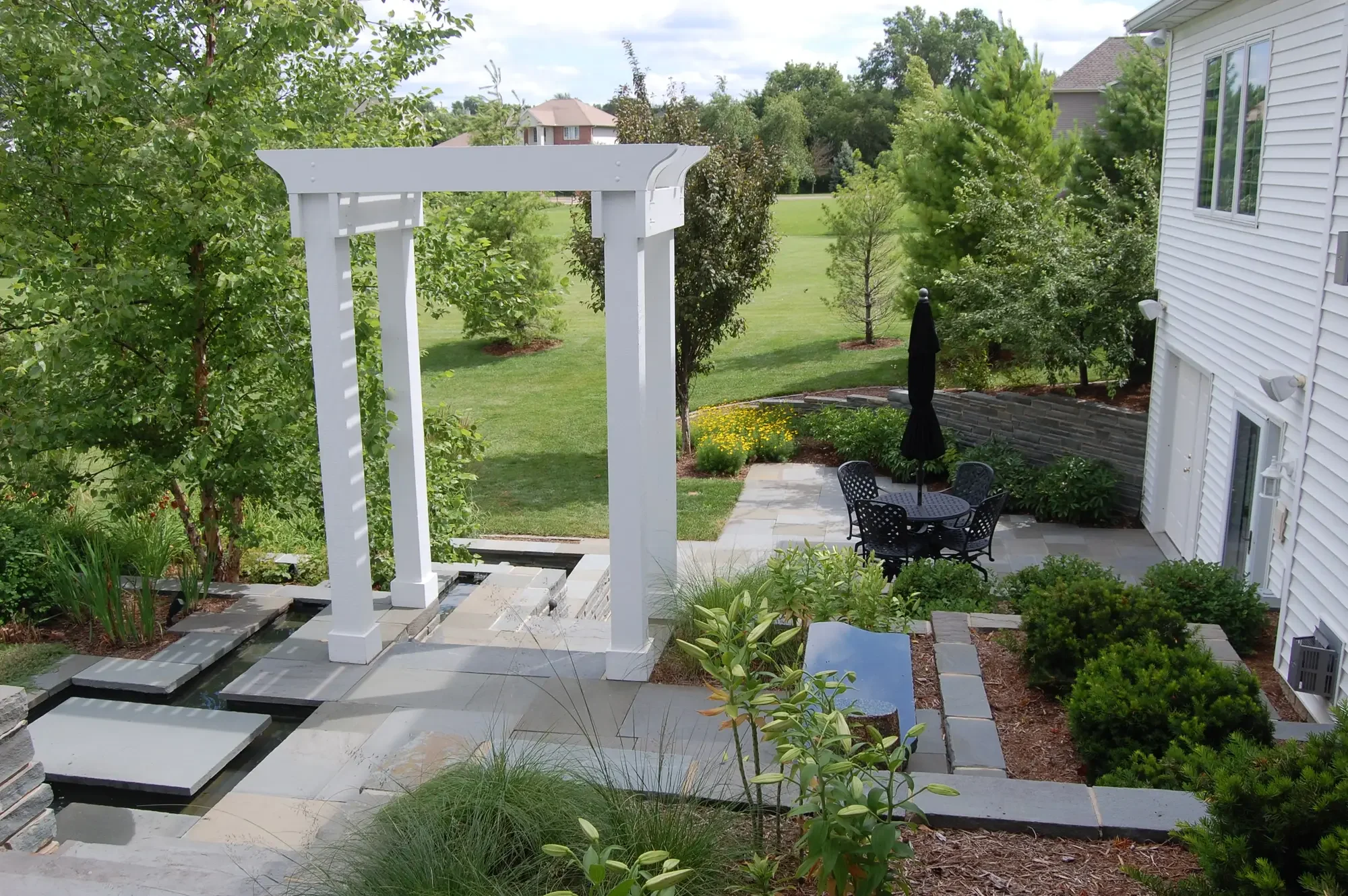 Elevated view of white pergola, floating bluestone water feature, patio dining area, and surrounding landscape at Harmony in Stone and Water by Green View in Illinois