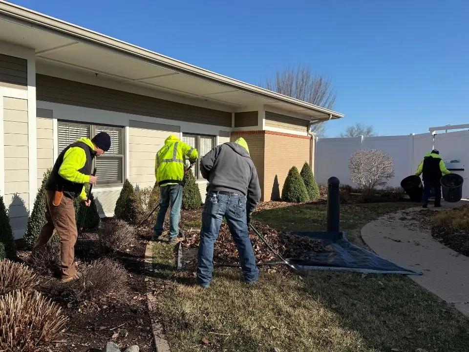 Four workers wearing safety vests and jackets removing mulch from a garden bed outside a house, with two the house's window and white fence in the background.