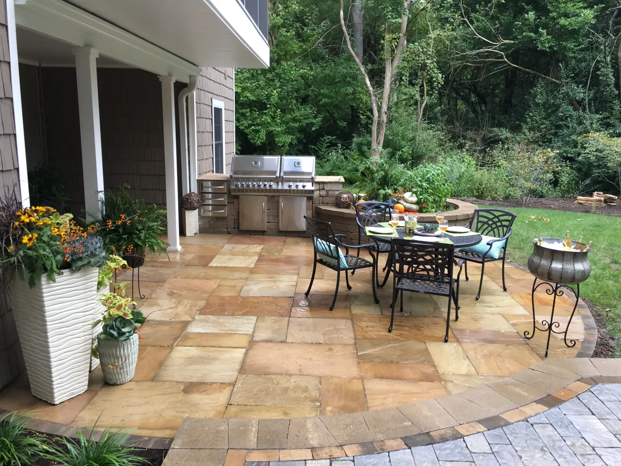 A backyard patio with a round dining table set for four, surrounded by four black metal chairs with blue cushions. To the left, potted plants with yellow flowers and greenery. In the background, a built-in stainless steel BBQ grill with a stone countertop, and lush green trees and grass beyond the patio.