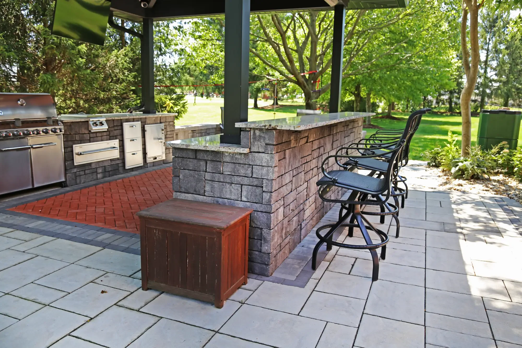 Outdoor kitchen with granite bar, bar seating, and herringbone brick paver floor inside pavilion at The Wooded Pavilion by Green View in Illinois
