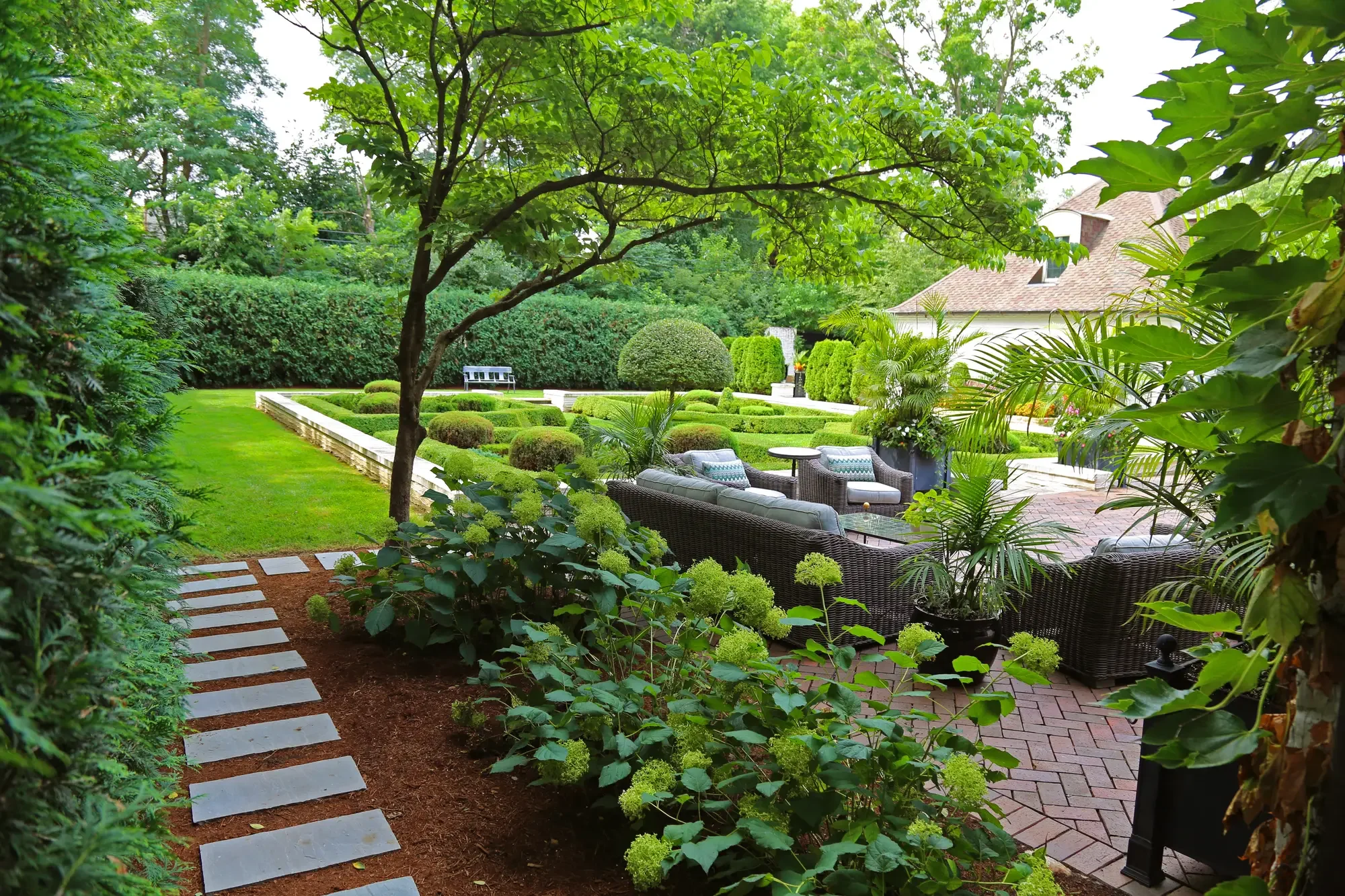 Garden view through hydrangeas and tropical plantings toward formal knot garden and brick patio seating area at The Tudor Knot Garden by Green View in Illinois