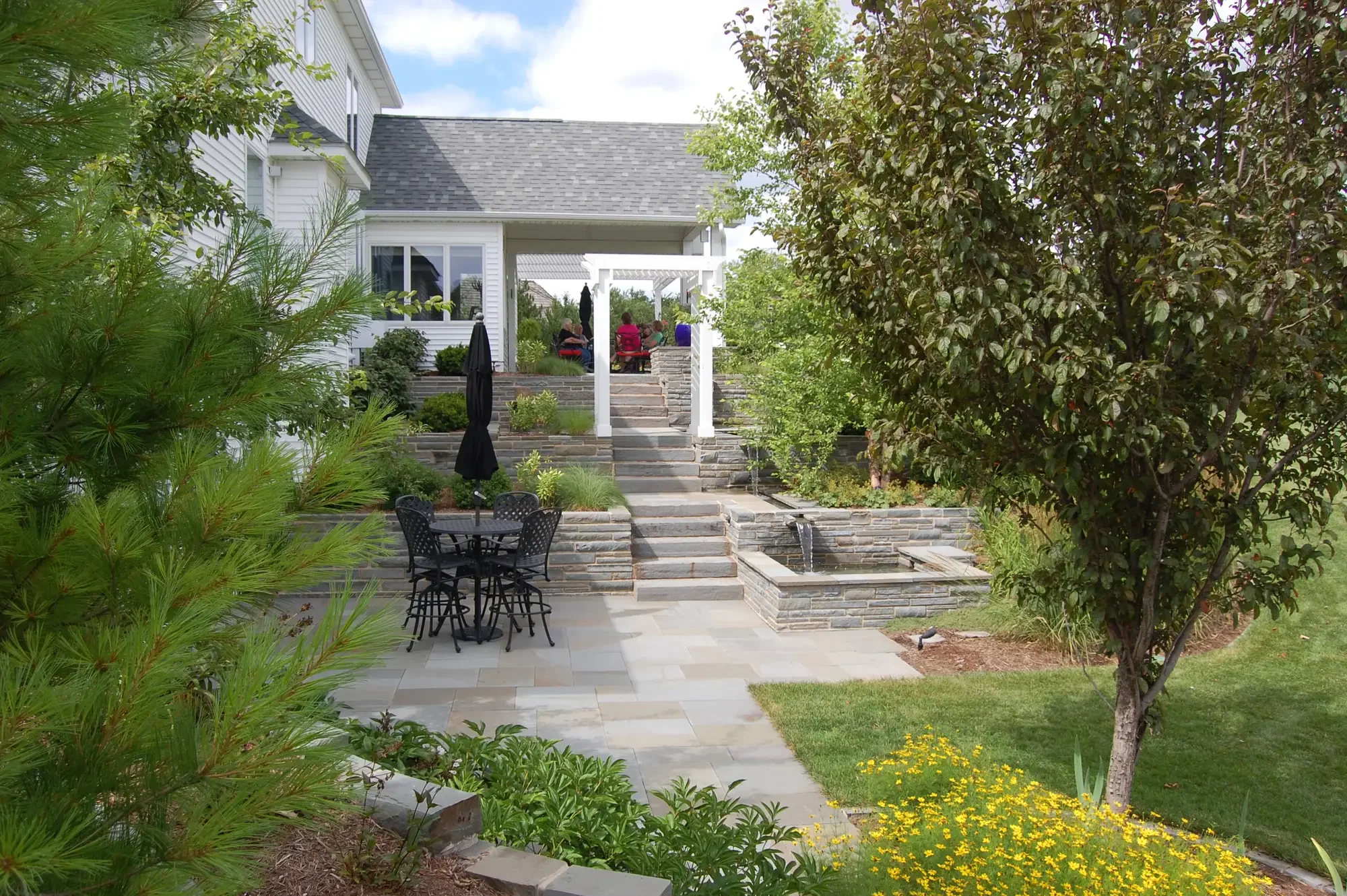 Multi-level bluestone patio with white pergola, stone water feature, and tiered stacked stone retaining walls at Harmony in Stone and Water by Green View  in Illinois