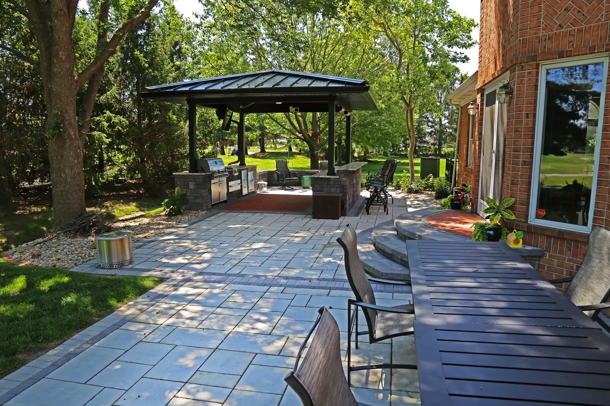Full backyard view of dark metal roof pavilion, large-format paver patio, and curved entry steps at The Wooded Pavilion by Green View in Illinois
