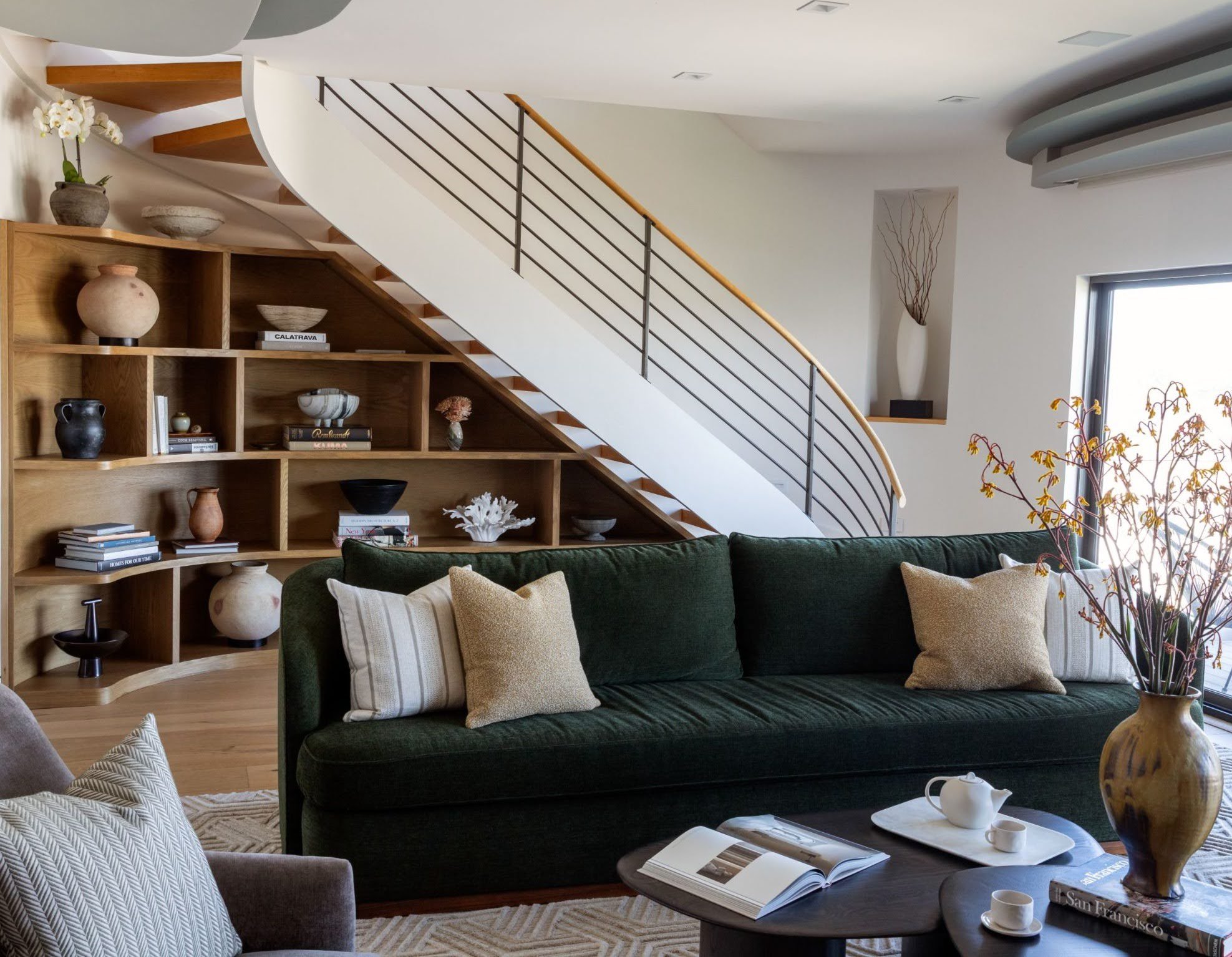 Living room with a deep green sofa, curved staircase with oak built-in shelving underneath, styled ceramics and a sculptural dark wood coffee table