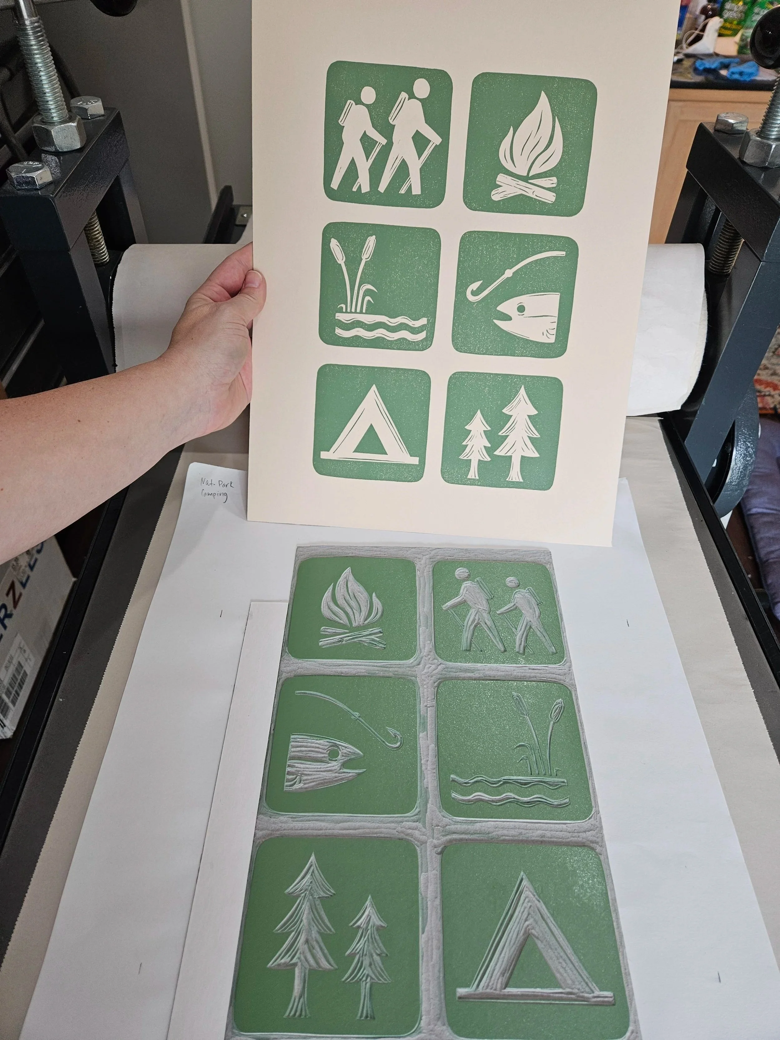 Process shot of hand carved linocut bring printed on paper featuring national park icons with outdoor adventure icons, including hikers, campfire, fishing, tent, trees, and nature elements.