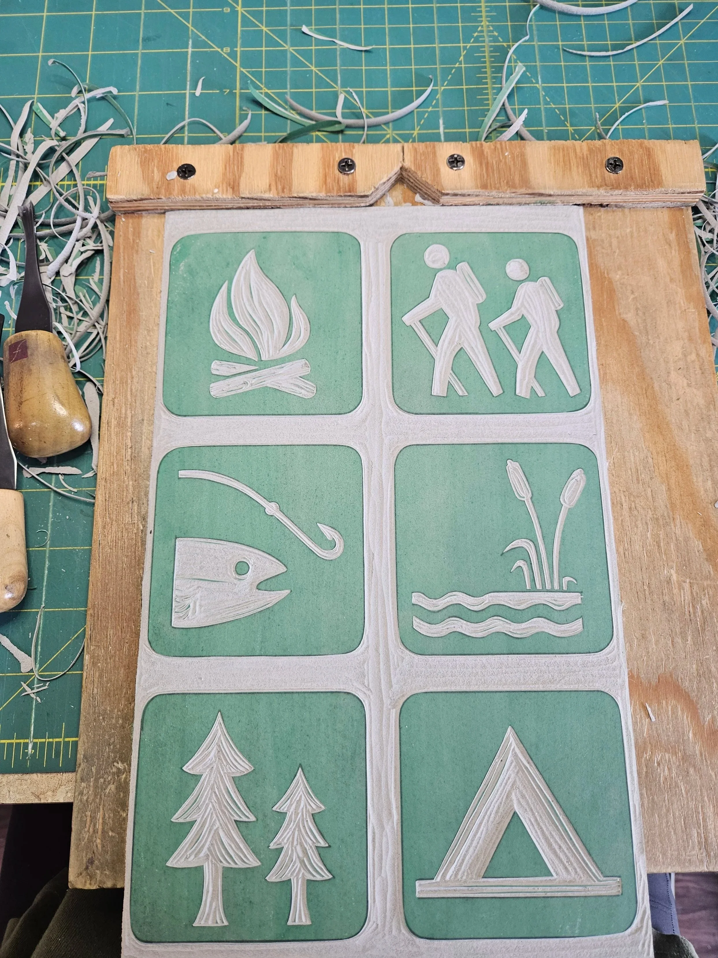 Hand carved linocut ready for printing on paper featuring camping and outdoor adventure icons, including hikers, campfire, fishing, tent, trees, and nature elements. National park inspired wall art.