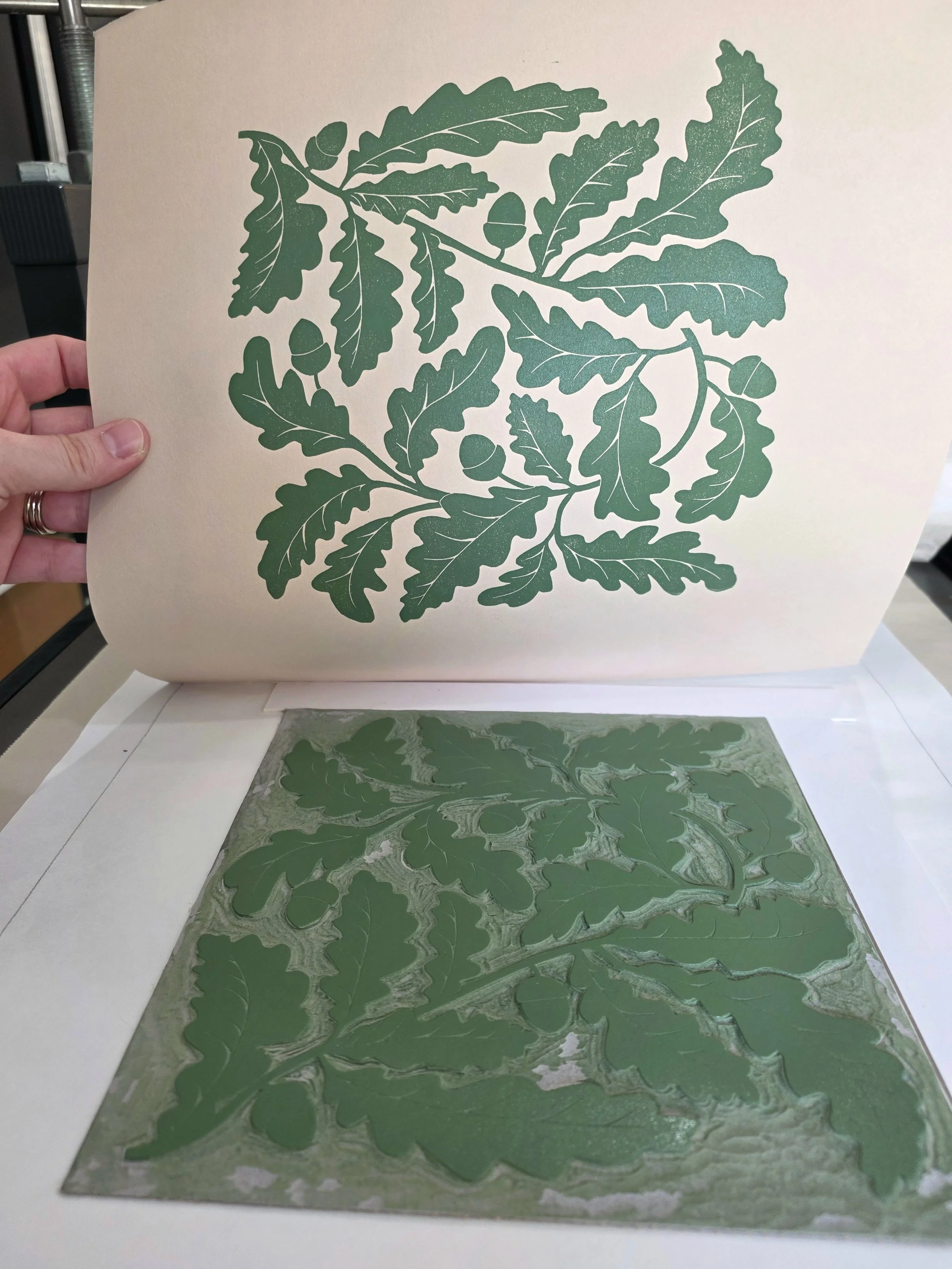 Handmade block printing process showing nature-inspired oak leaves and acorns design being lifted from the carved block, with olive green ink printed onto beige fine art paper.