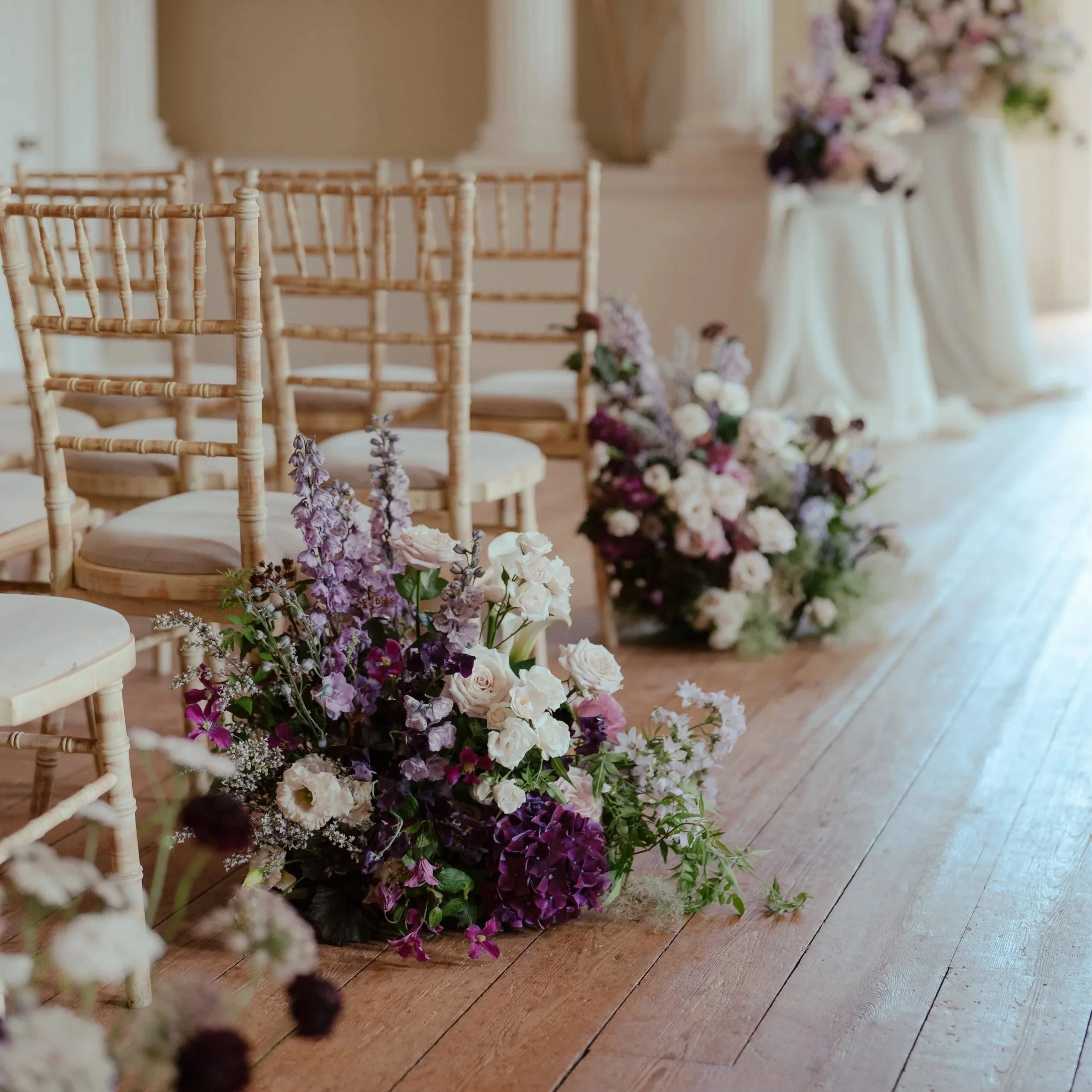 A wedding set in the county of Norfolk and Suffolk. Kimberley Hall. Wedding aisle decorated with floral arrangements on the floor and chairs, set in an elegant room with large windows, columns, and a portrait on the wall.