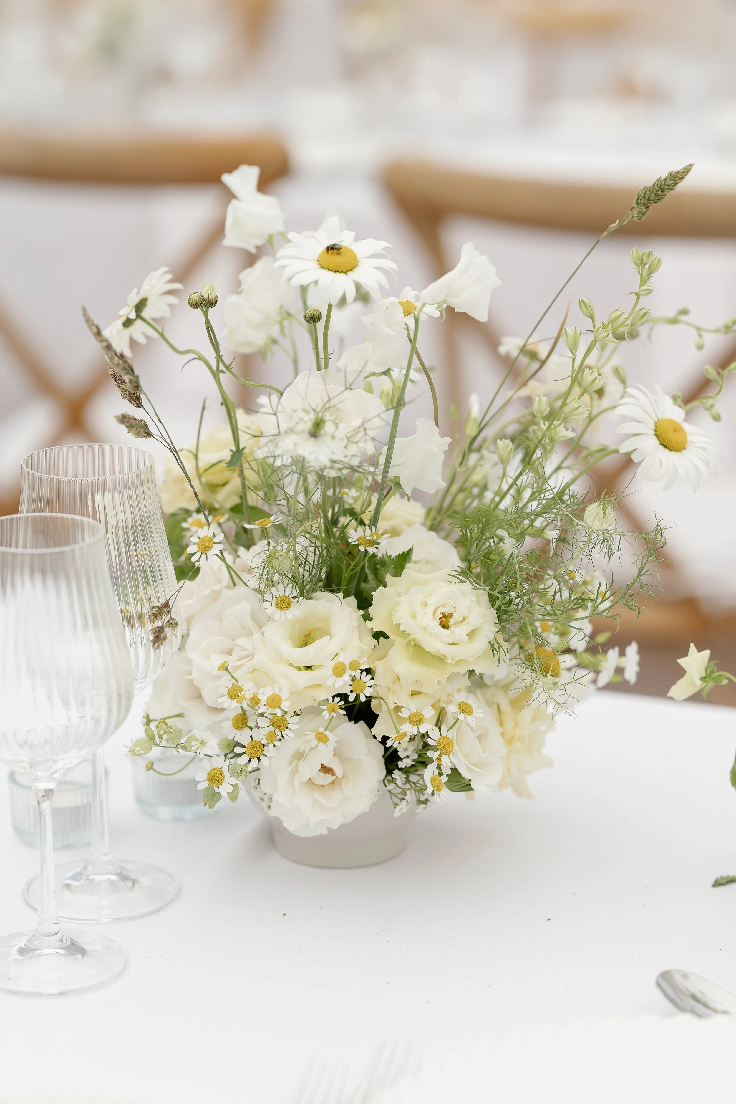 White, chic and luxury floral centrepiece with daisies, roses, and greenery on a table with wine glasses. Taken by Norfolk's best wedding photographer Sharon Cudsworth. 