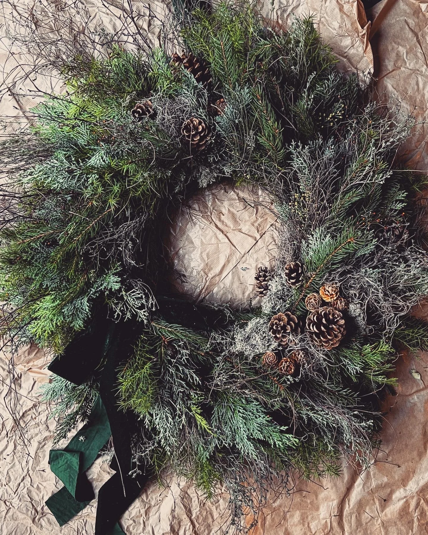 A decorative Christmas wreath made of various evergreen branches, pinecones, and frosted foliage, arranged on crumpled brown paper. Created with our christmas wreath workshops at a North Norfolk location
