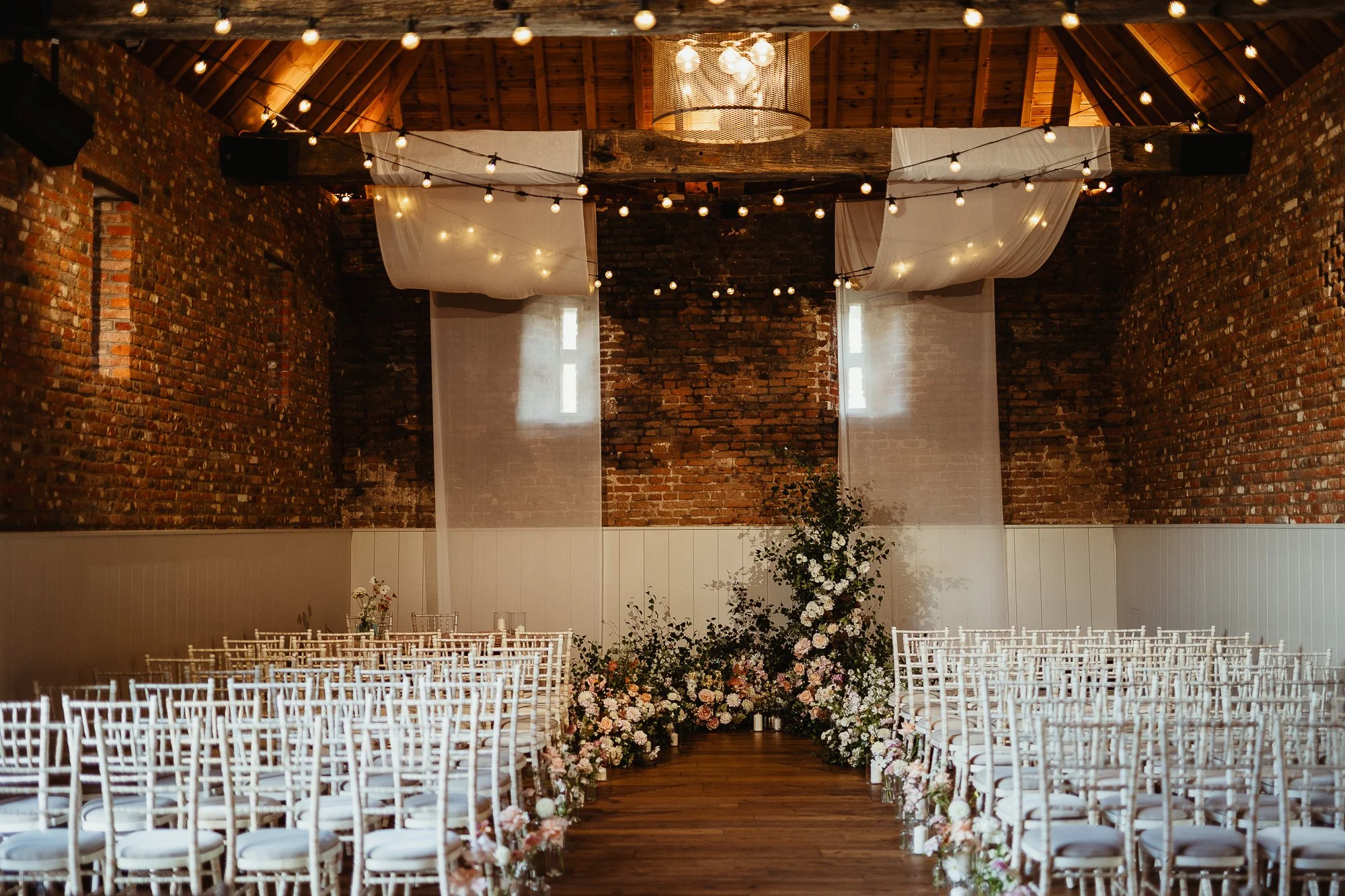 Indoor wedding ceremony setup with white chairs lining a wooden aisle, floral deconstructed arch at the end, exposed brick walls of Worstead Barn in Norfolk, string lights, and draped fabric on the ceiling.