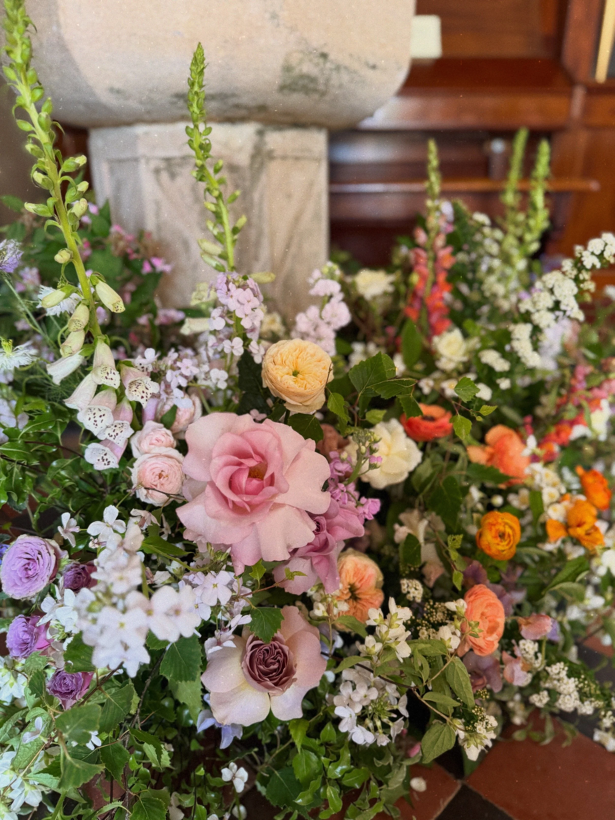 Assorted colorful seasonal wedding flowers including pink roses, purple ranunculus, white and pink snapdragons, and orange marigolds, arranged in a lush floral display in Oxnead Hall Church in Norfolk