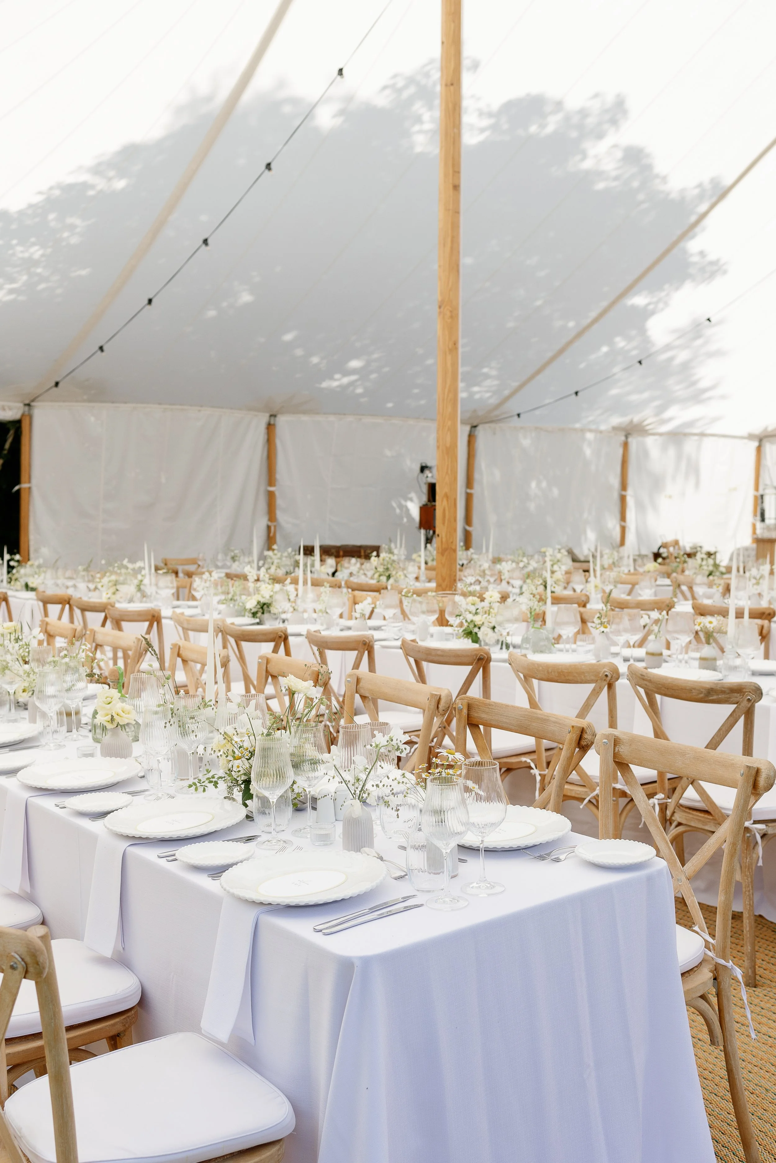 Elegant and luxury event setup with long white-draped tables, wooden chairs, and compote / bowl floral centrepieces under a white tent by Sail and Peg. 