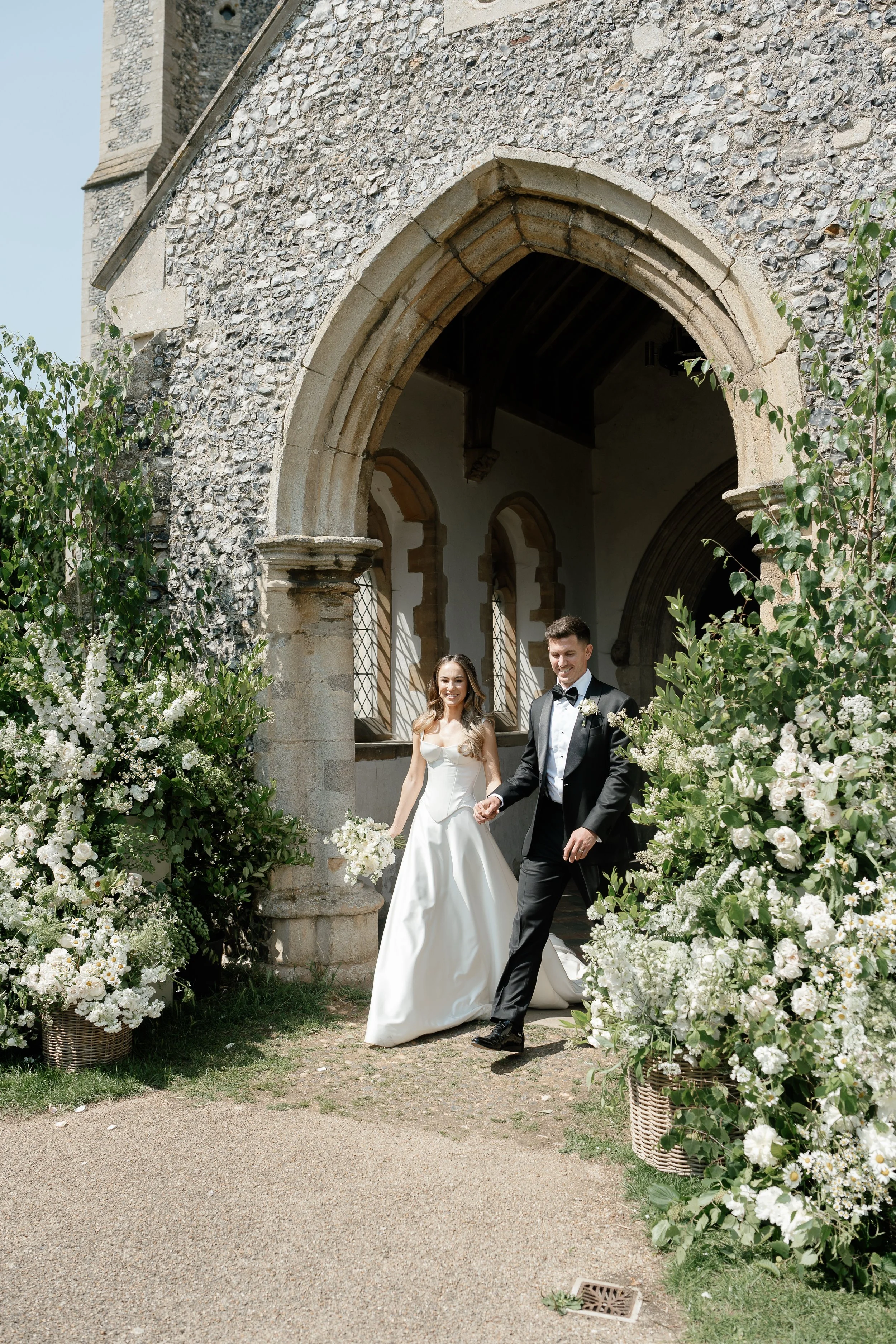 A bride and groom walking out of a historic stone church in Norfolk, holding hands, with lush white floral arrangements surrounding them, on their wedding day.