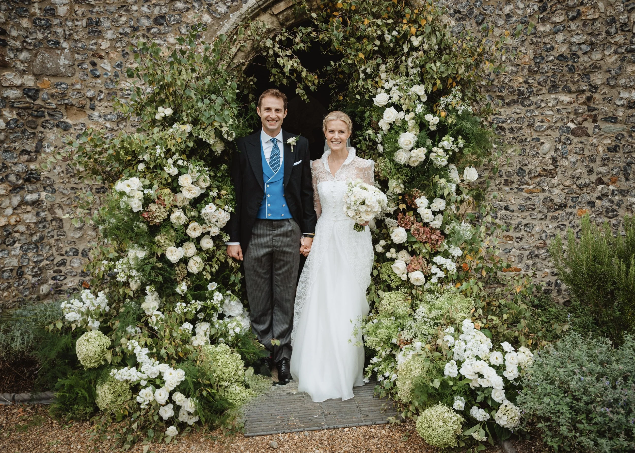 A bride and groom holding hands, standing under a floral archway made of white and green flowers, in front of a Church in Ingham, Norfolk. Created by the best wedding florist in Norfolk and Suffolk.
