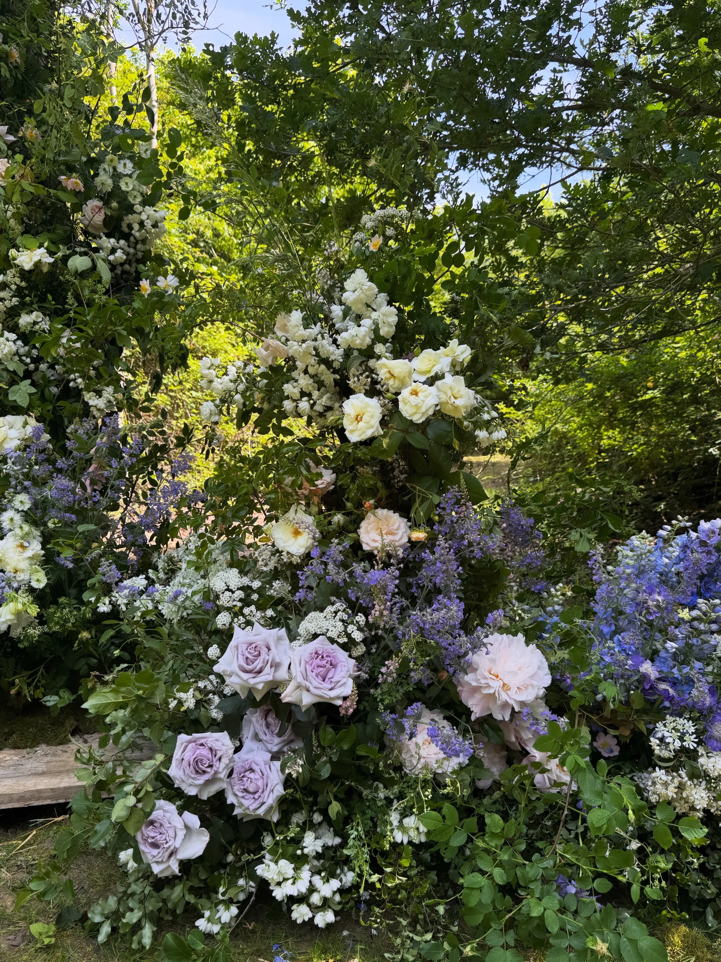 A lush woodland ceremony in Norfolk farmland, covered in garden-type white and purple flowers, including roses and other Peonies, surrounded by wild green foliage.