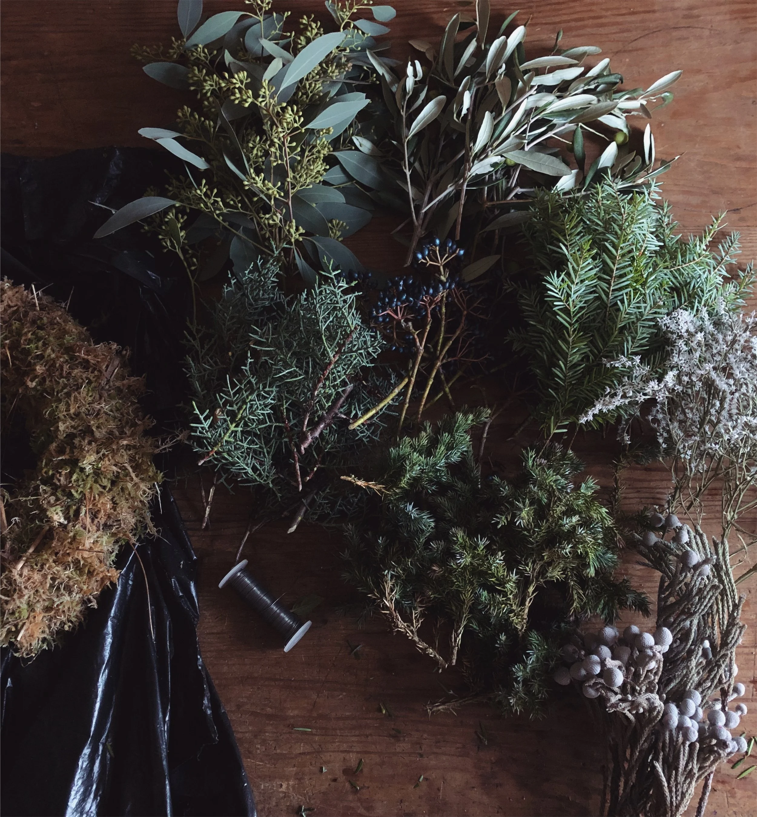 Various types of greenery, including sprigs of eucalyptus, pine, and other foliage, arranged on a wooden surface with some decorative white balls at the bottom right. An example of what a work station will look like at our festive wreath making class