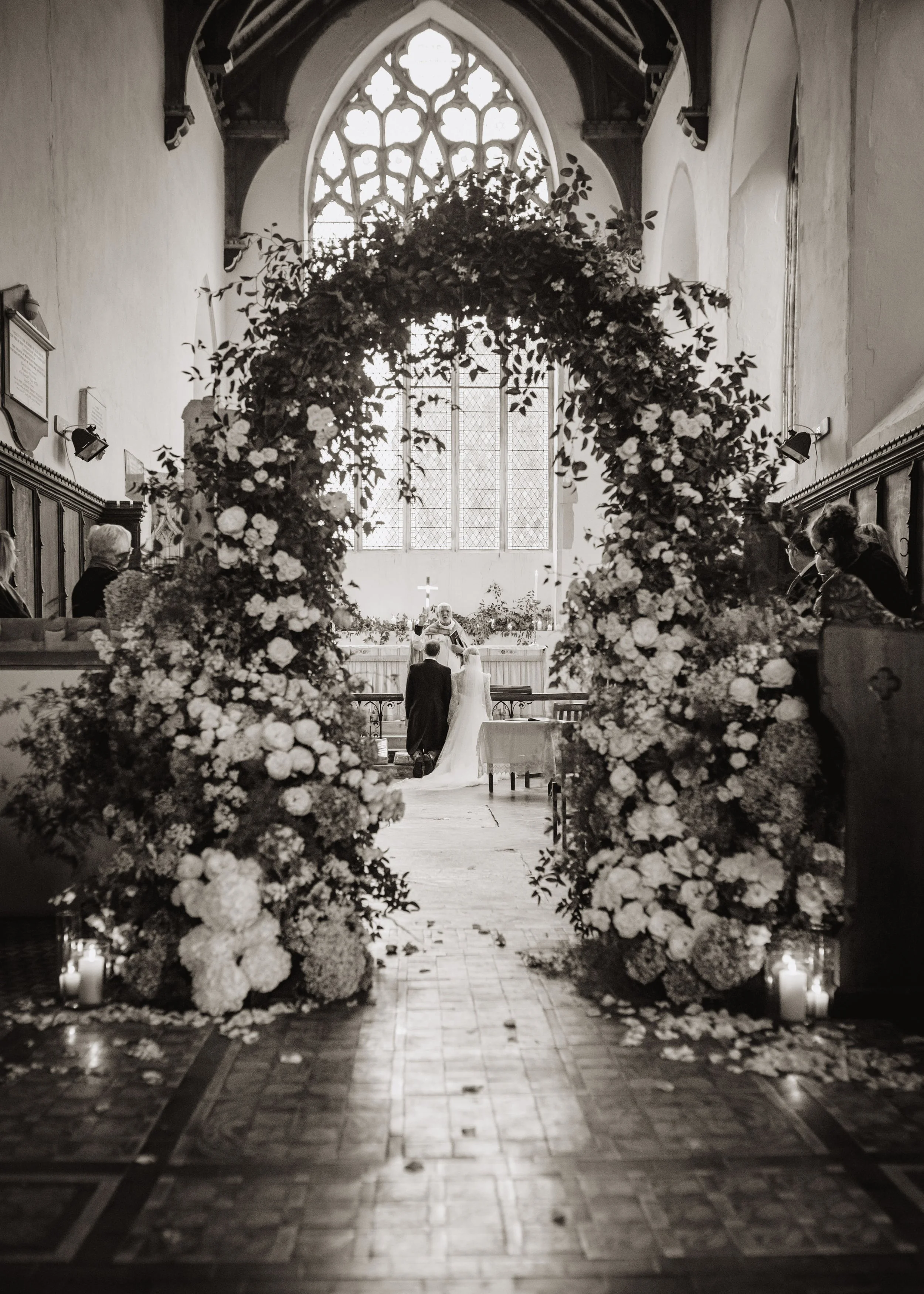 A black and white photo of a wedding ceremony inside a church with a large floral romantic archway and stained glass window. Only the best florist up Norfolk and Suffolk created this design