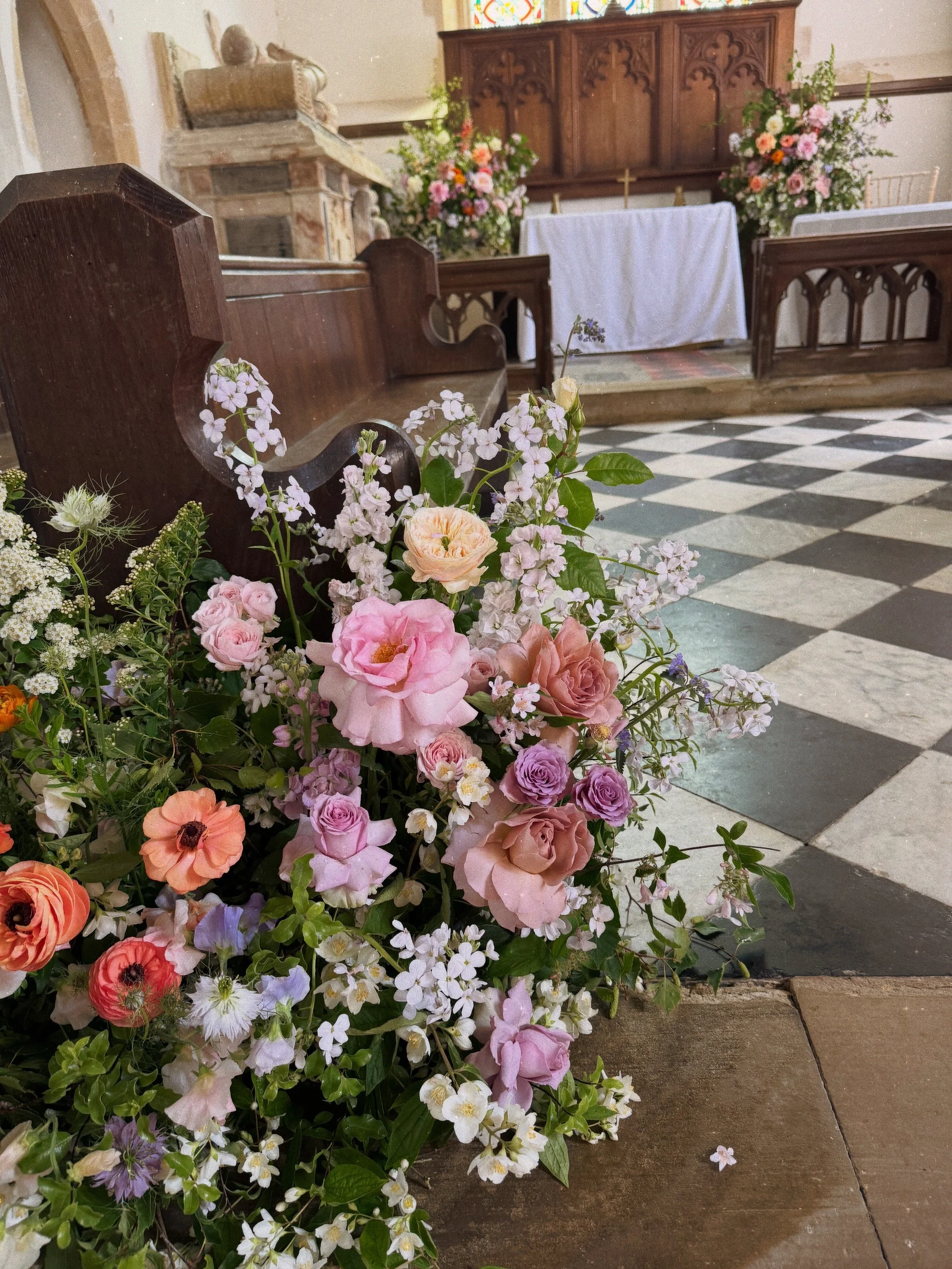 Oxnead church in Norfolk, the church interior with wooden pews, a stone pulpit, and floral arrangements including pink, purple, and white flowers near the altar, which is covered with a white cloth and has a wooden backdrop with a cross.