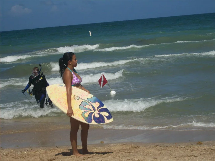 Photo of the author holding a skim board at Lauderdale by the Sea while a scuba diver comes out of the waves. Ancestral land of the Seminole tribe.