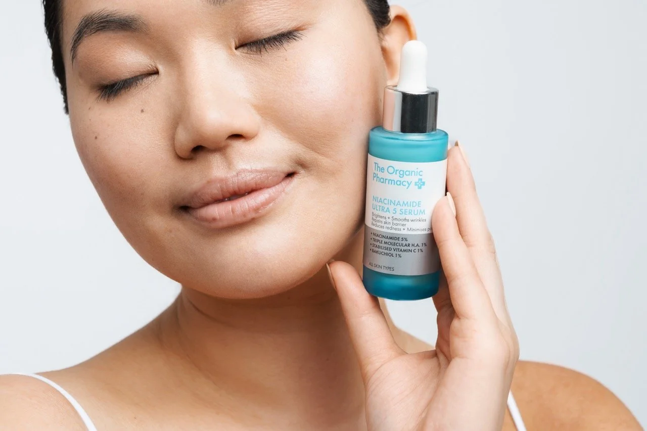 A woman with closed eyes holding a blue bottle of skincare serum labeled 'Niacinamide Ultra 5 Serum' from The Organic Pharmacy, showing the product close to her face.