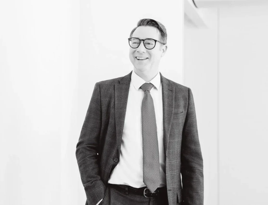 A smiling man in a suit with glasses standing indoors. CEO headshot for updated LinkedIn and press release images. Corporate headshots