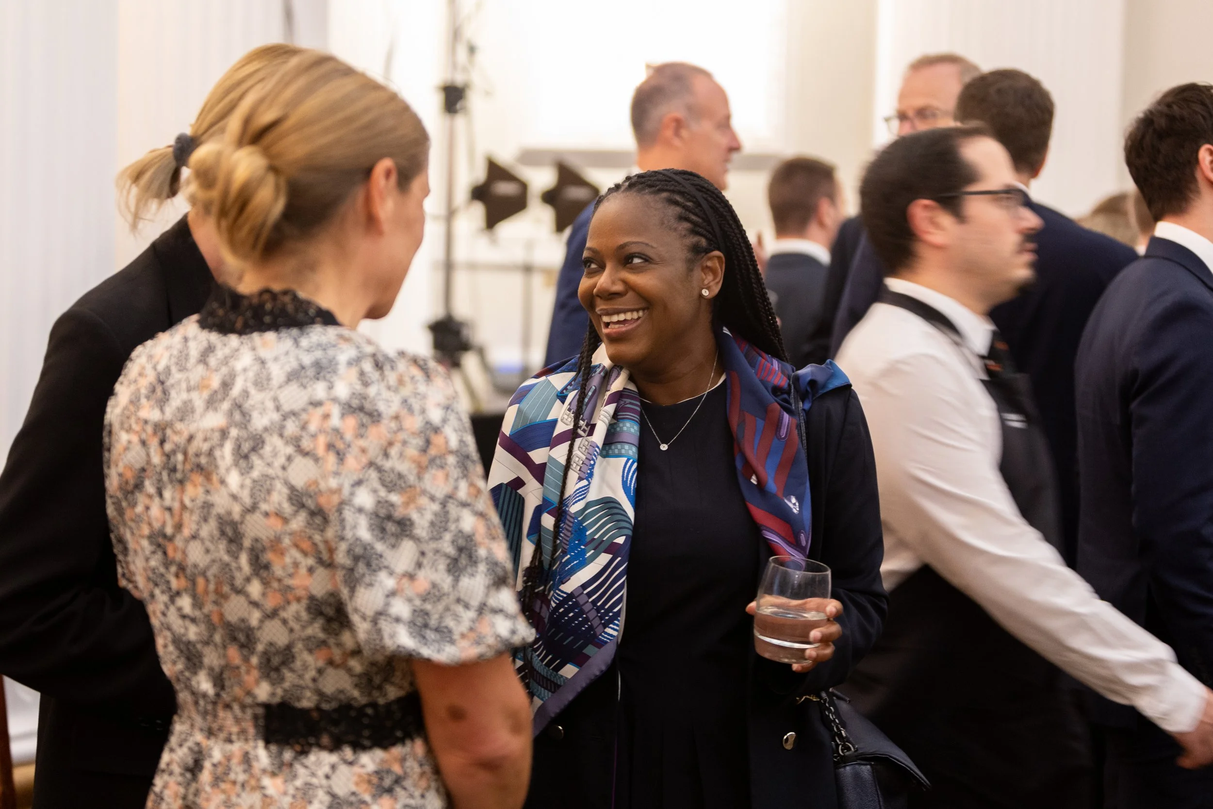A woman with braided hair wearing a colorful scarf and black top, smiling and holding a glass, engages in conversation with two women at a formal gathering.