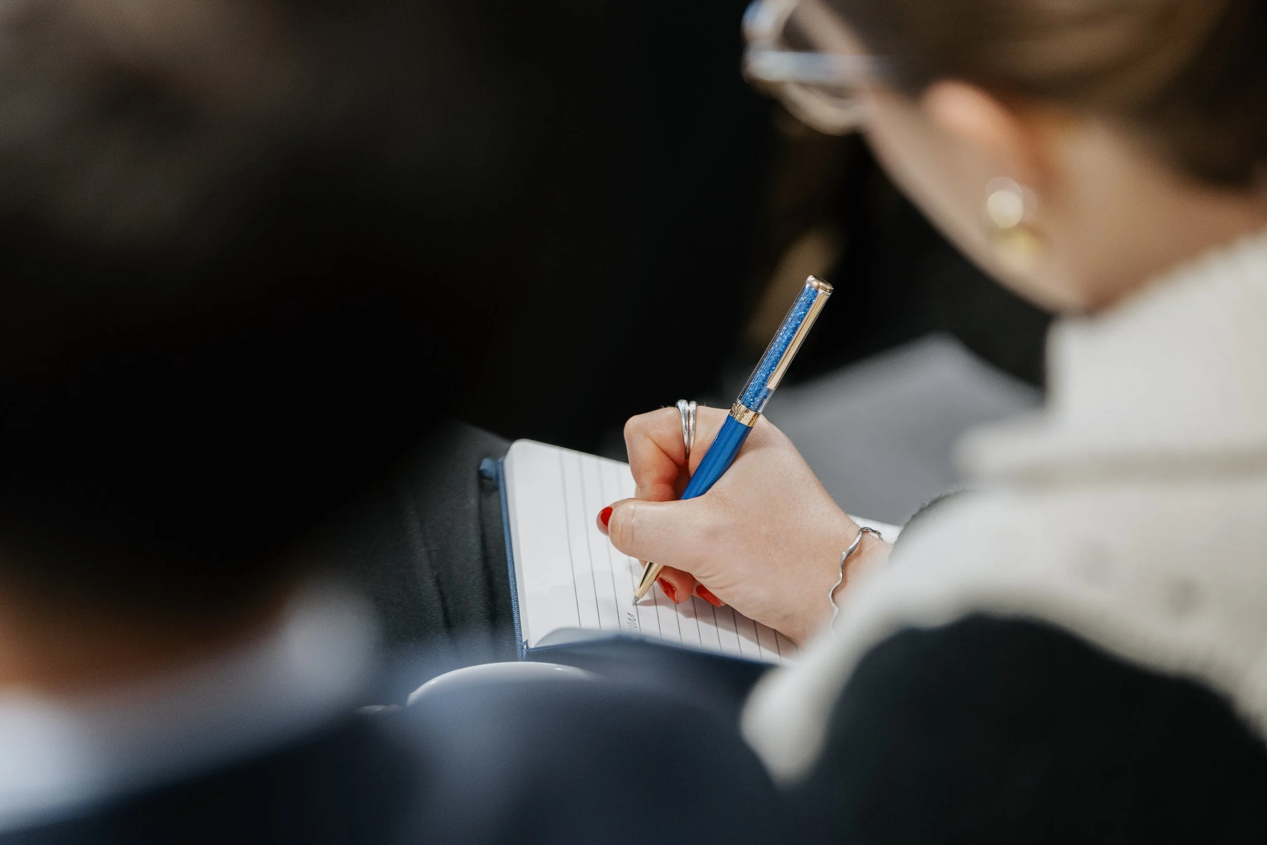 A woman with jewelry, wearing glasses, writing in a notebook with a blue pen during a meeting or conference. Corporate Photography, event photography