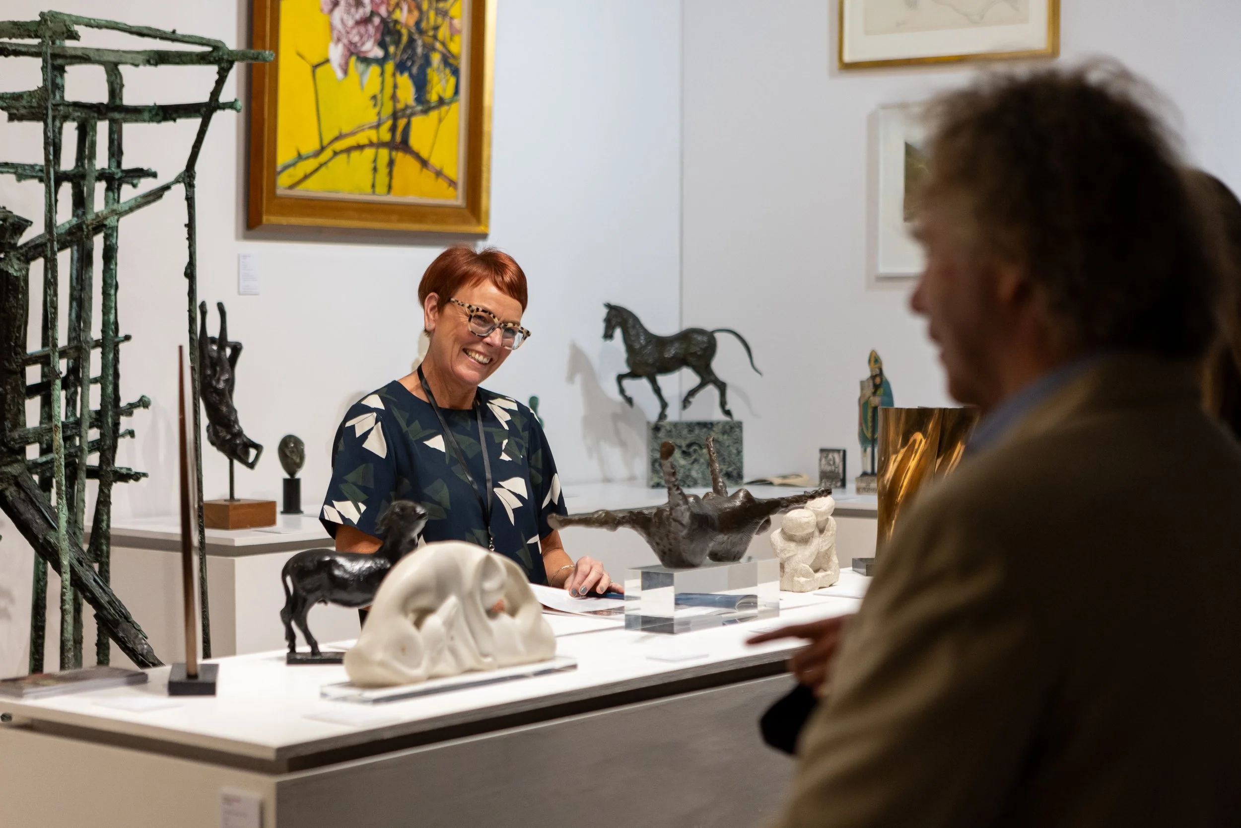 A woman with short red hair, glasses, and a blue patterned shirt looks at a visitor in an art gallery. The gallery display includes sculptures of animals and skulls, with paintings on the wall behind.