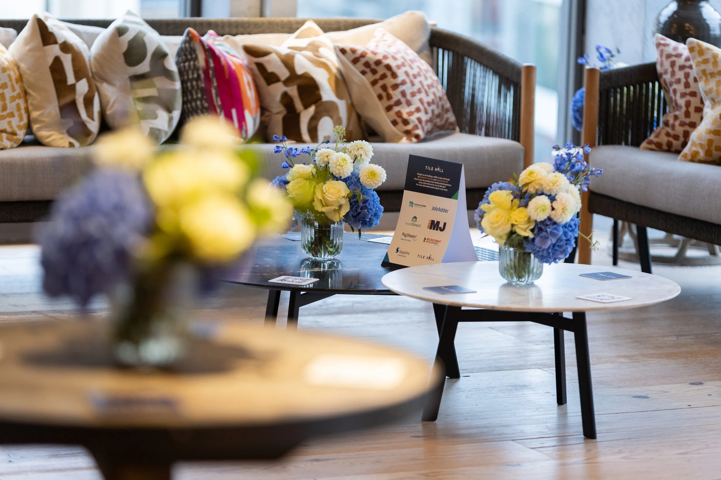 Two small round tables with flower arrangements, and a sofa and chairs with cushions in a bright indoor space with large windows. Tile Hill Recruitment summer event with networking and headshots. 