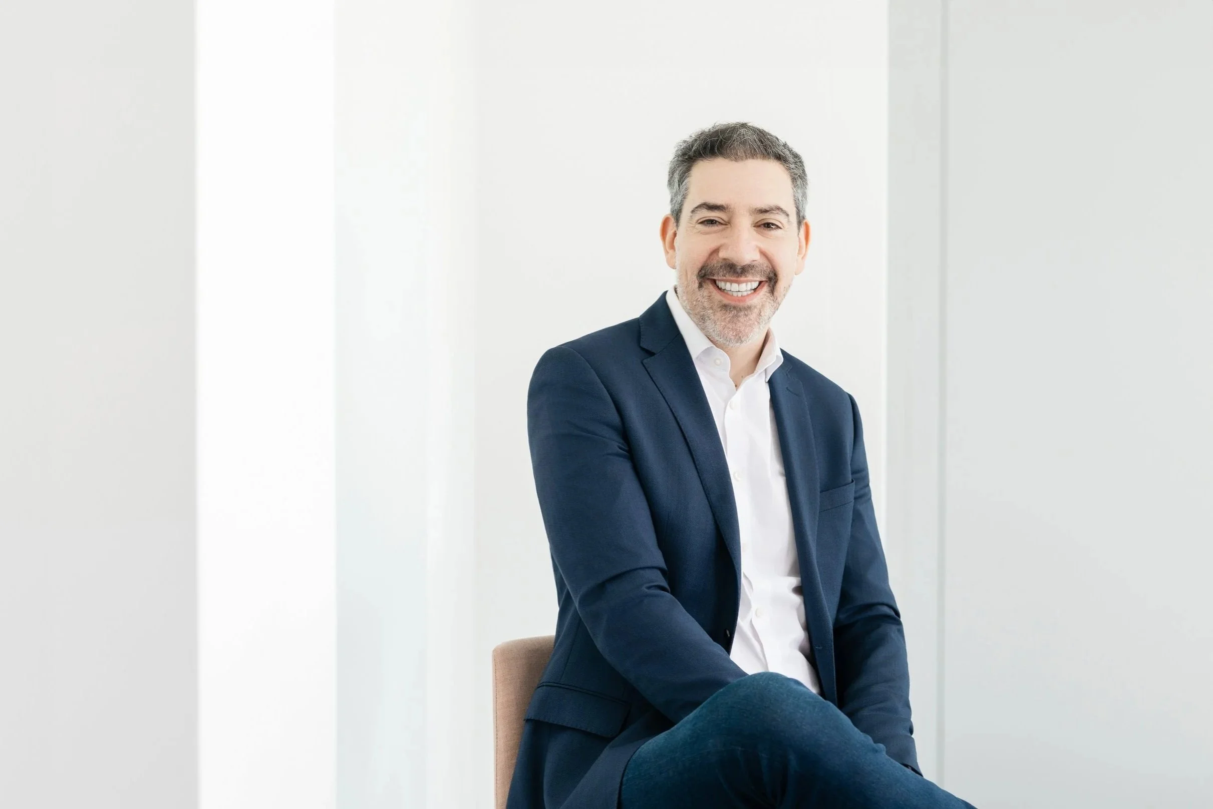 A smiling man with gray hair and beard wearing a navy blazer and white shirt, sitting on a beige chair against a white background. Corporate headshot photography.