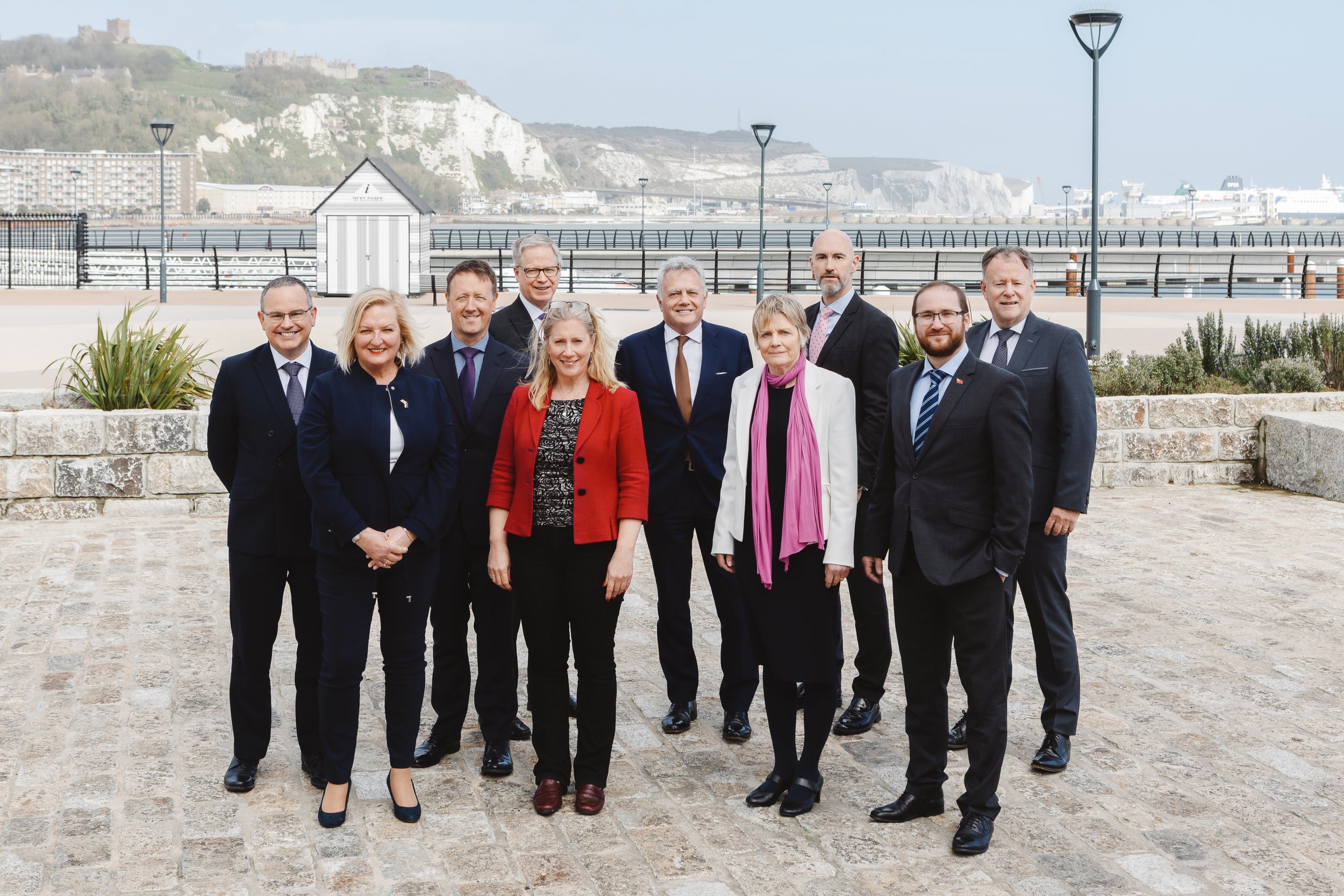 Dover Ports group corporate headshots including men and women, standing outdoors on a stone-paved area with a coastal background featuring cliffs, a harbor, and a cruise ship. The group is smiling and posing for the photo.