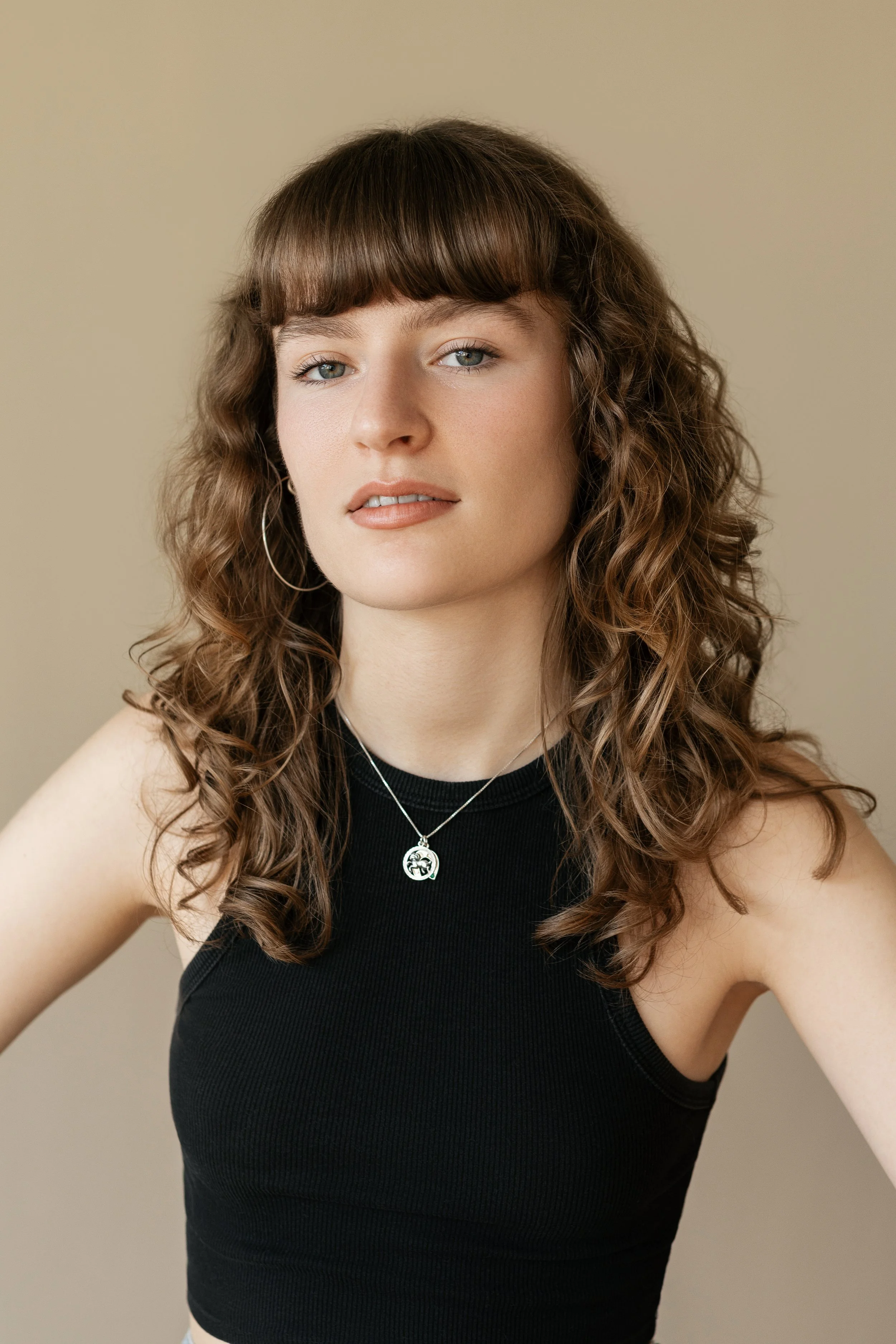 A young woman with curly brown hair, blue eyes, wearing a black sleeveless top, hoop earrings, and a necklace with a pendant, posing against a beige background. Actor headshots 
