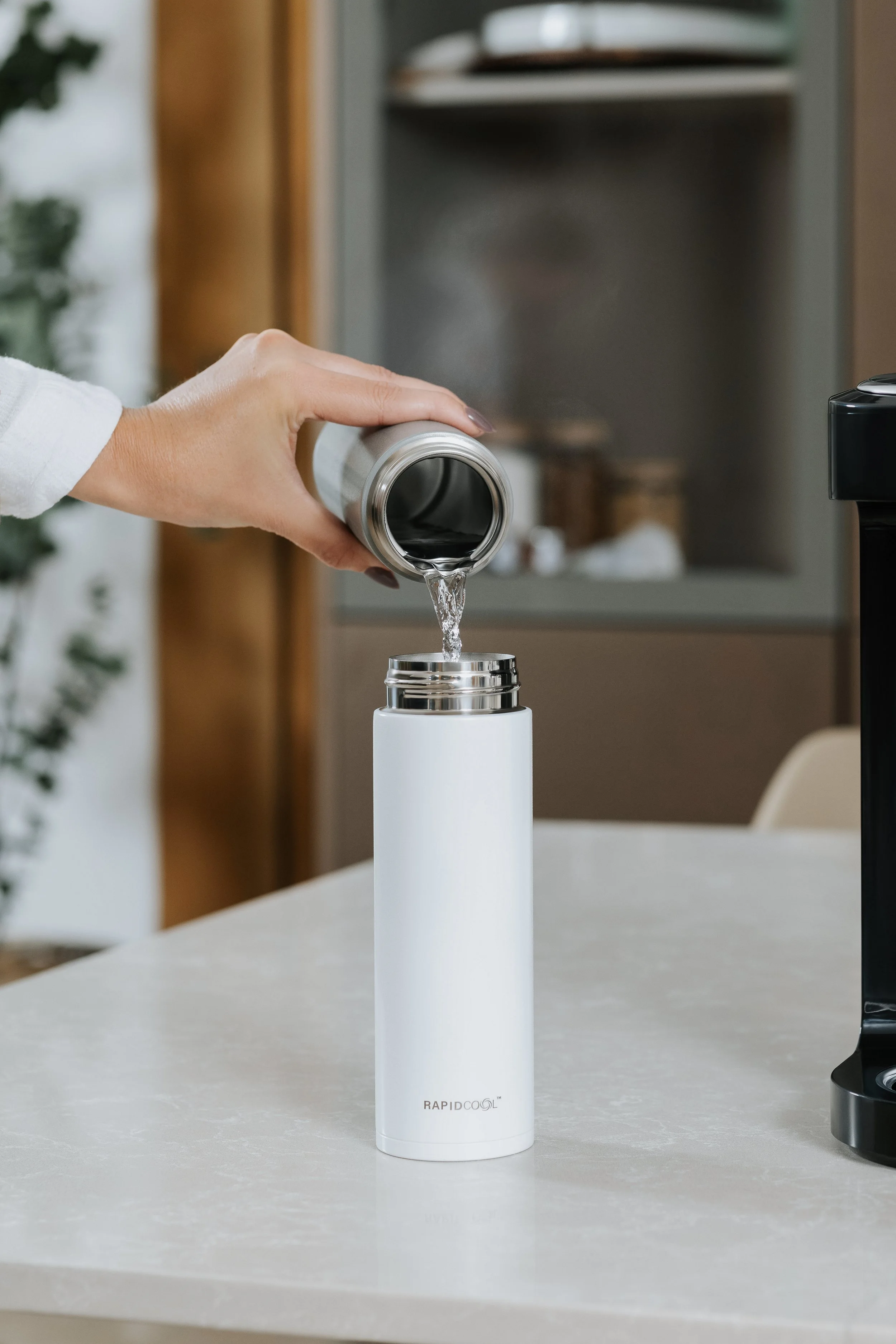 Person pouring liquid into a white insulated water bottle in a kitchen. Commercial lifestyle content for baby brand Nuby