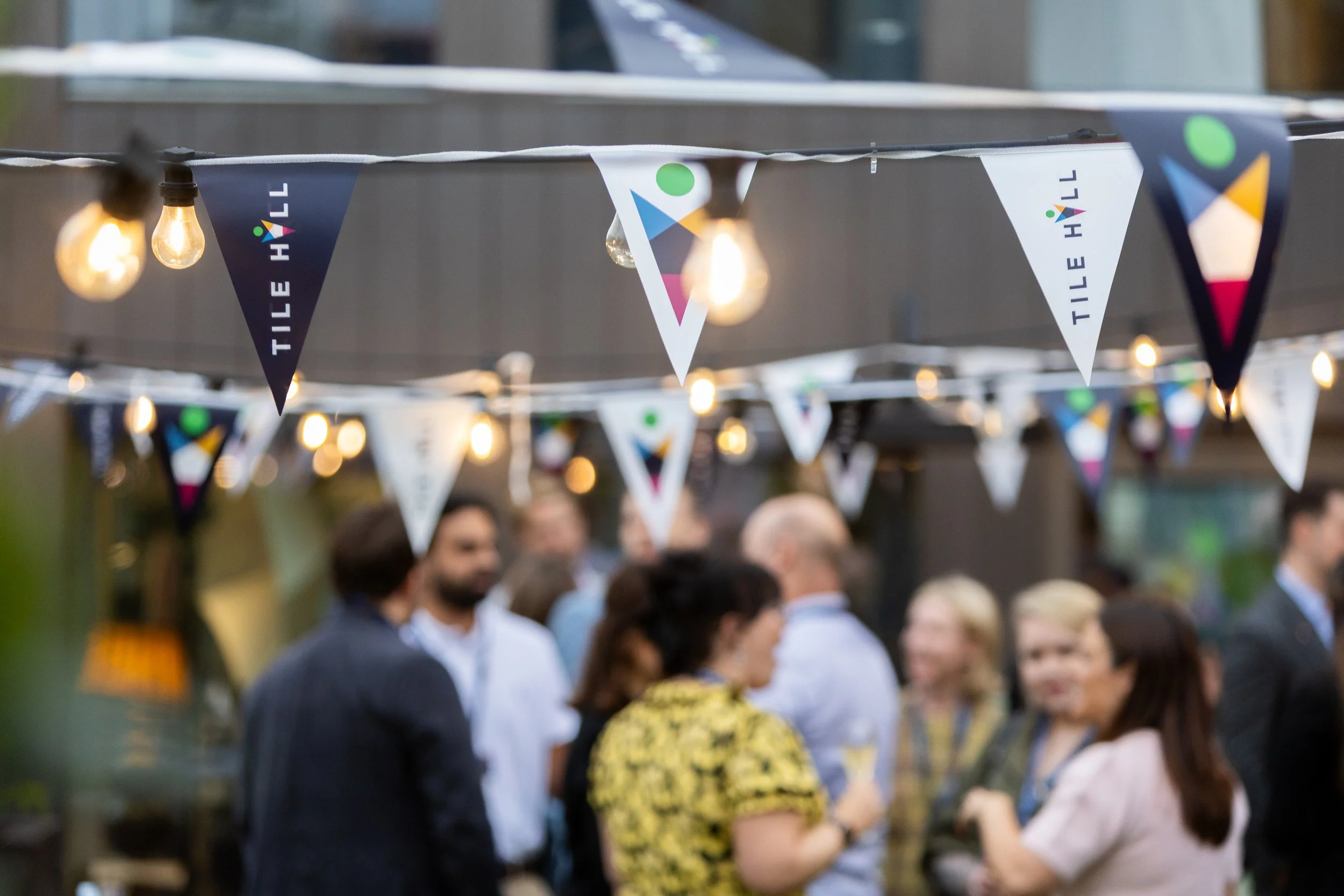 People socializing at an outdoor event with string lights and decorative banners reading Tile Hill - corporate evening event 