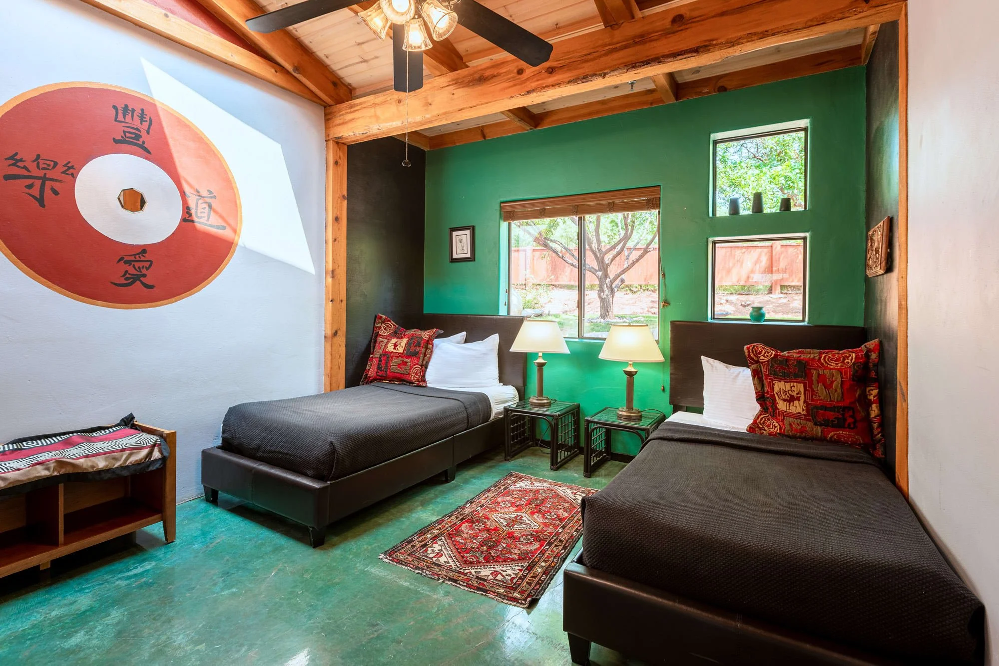 A bedroom with two twin beds, each with black bedding, red decorative pillows, and white pillows. Between the beds are two bedside tables with lamps. The room has a green accent wall with windows, a wooden ceiling with exposed beams, and a ceiling fan. There is a colorful rug on the green floor and a large wall mural featuring a red circular art piece with black Chinese characters.