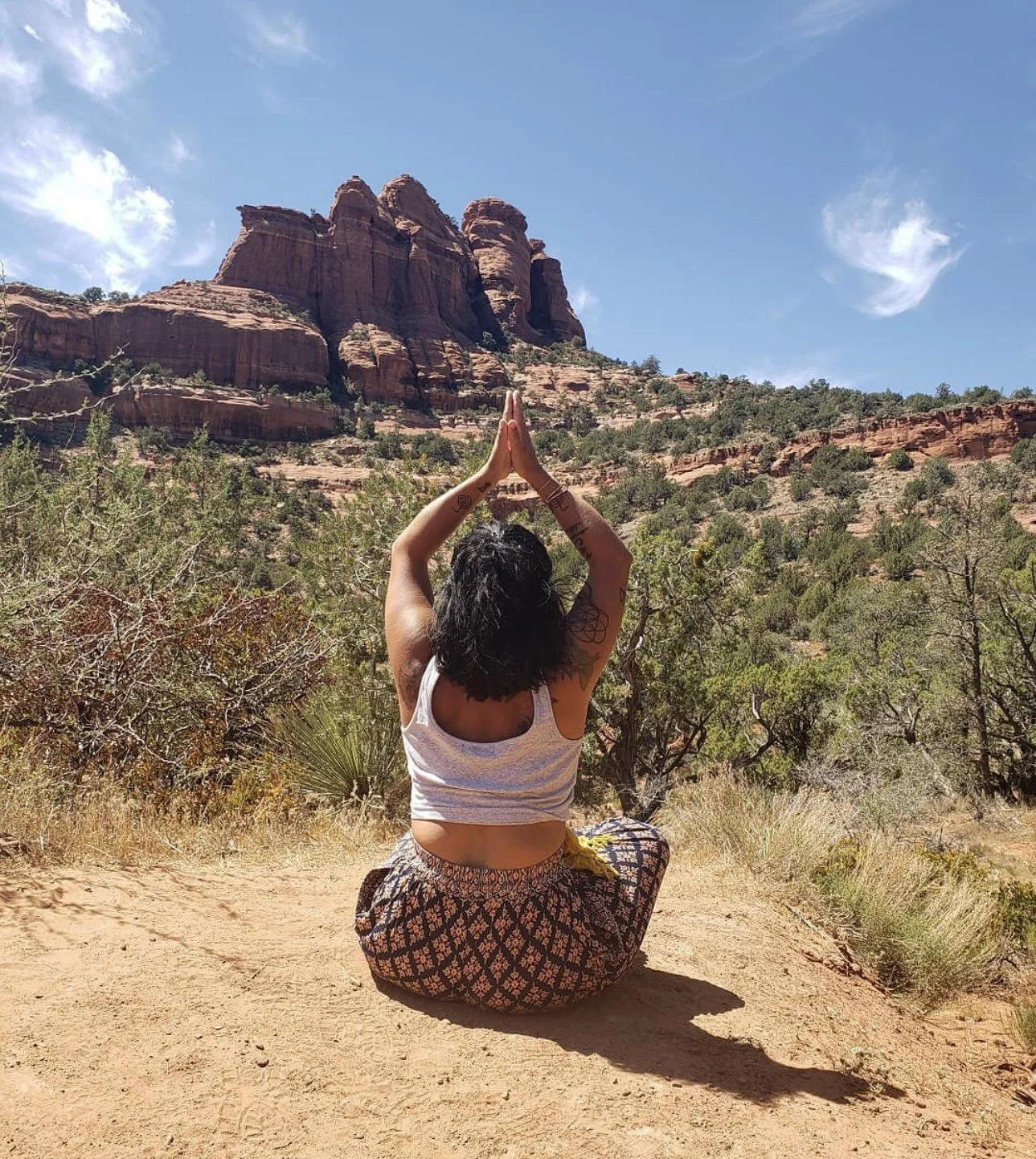 A woman practicing yoga in a desert landscape with red rock formations in the background, sitting cross-legged with hands raised in a prayer position.