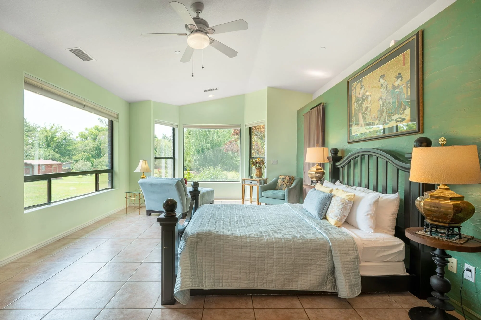 Bright bedroom with large windows, a bed with white bedding, blue and beige pillows, two bedside lamps, a green accent wall with a framed artwork, a small sitting area with chairs, a ceiling fan, and tiled floor.