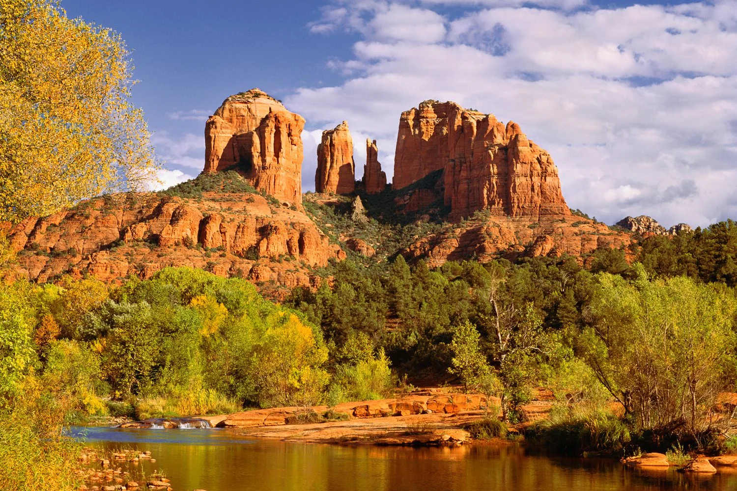 Red rock formations in Sedona, Arizona, with a river, green trees, and a partly cloudy sky.