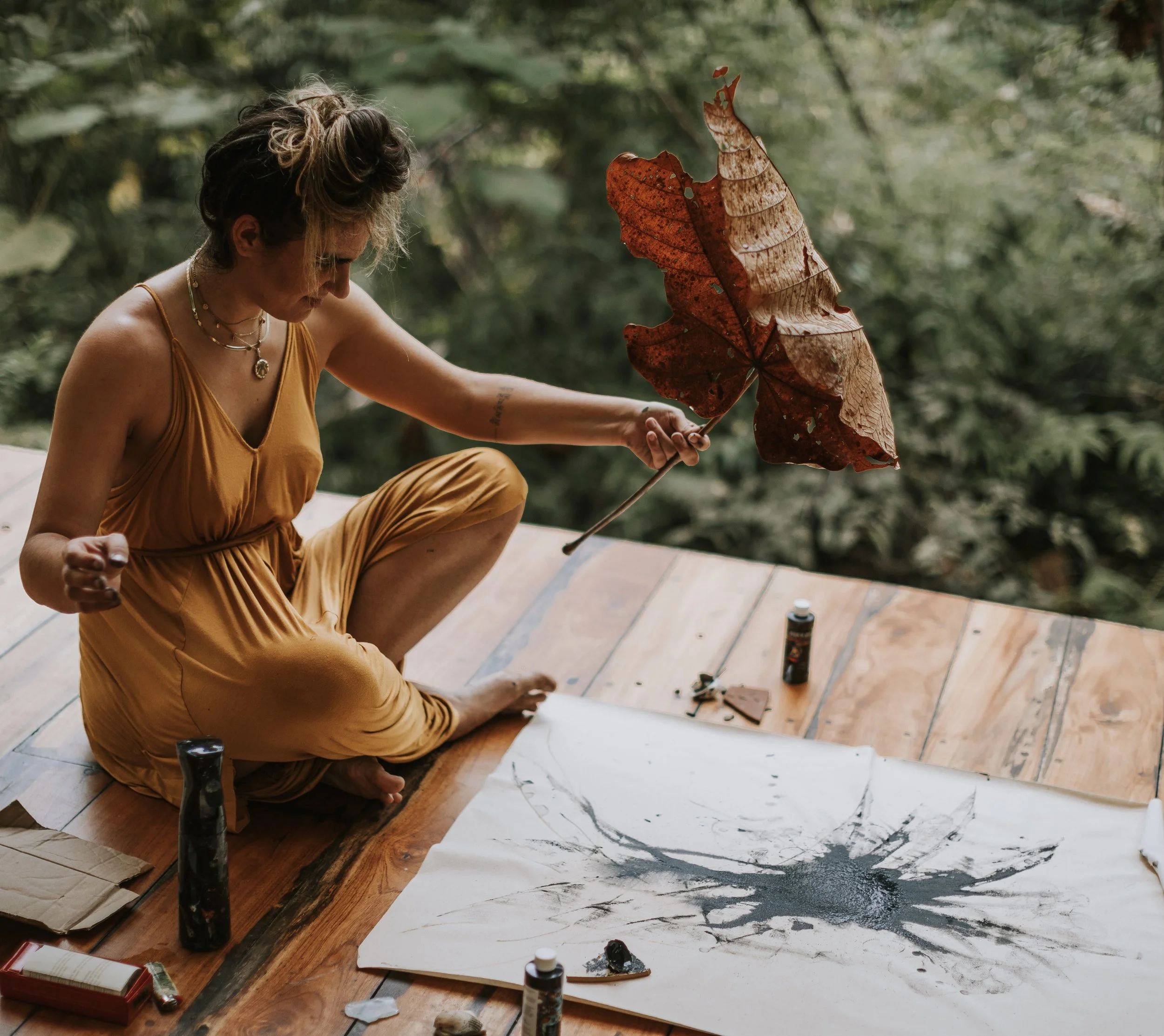 A woman in a yellow dress sitting on a wooden floor, holding a large brown dried leaf and creating an ink painting on paper with black ink splatters, surrounded by art supplies in a lush green outdoor setting.