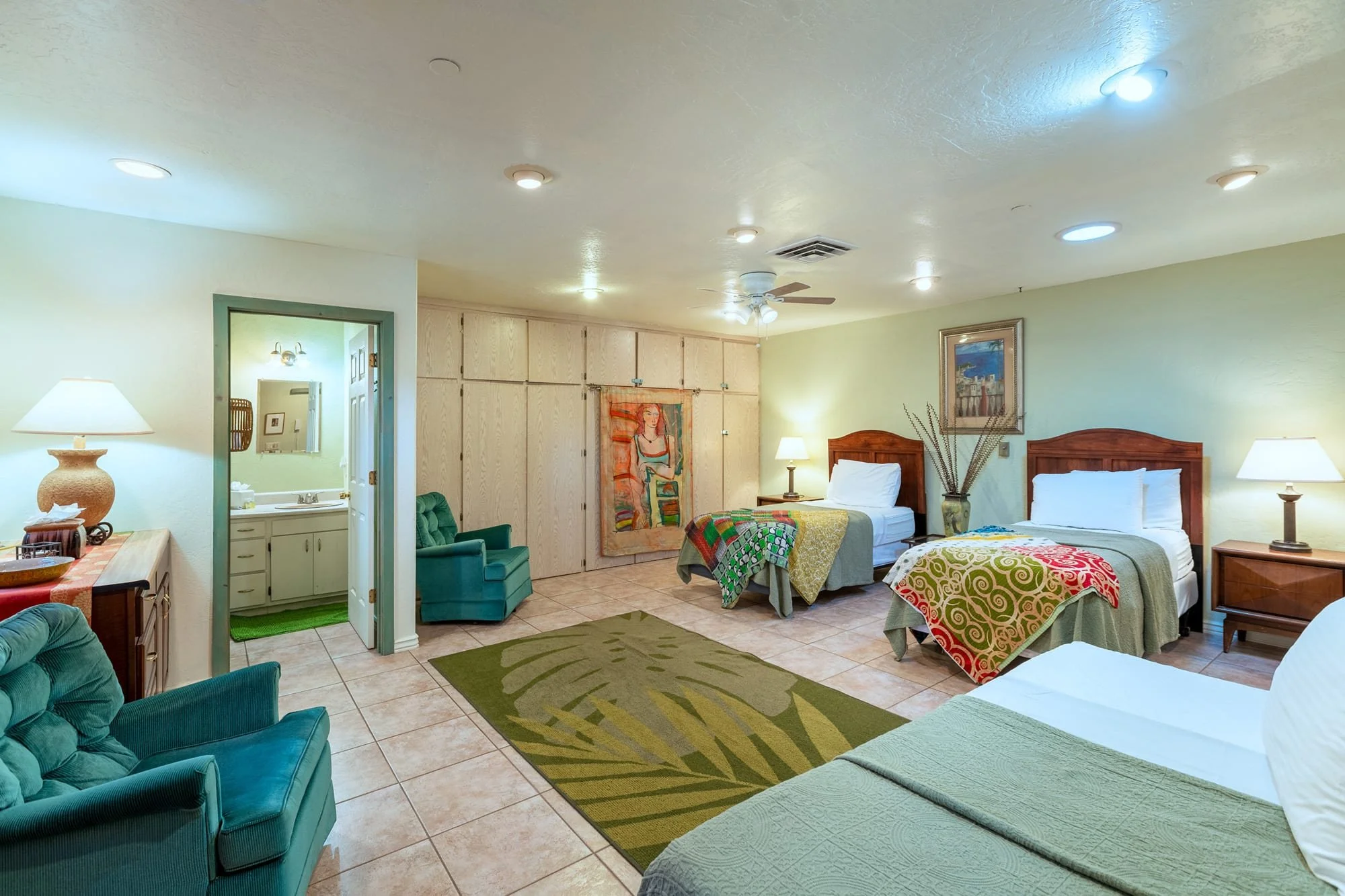 Interior of a hotel room with four beds, two nightstands with lamps, a green armchair, and a bathroom visible through an open door. The room has tiled flooring, a colorful rug, and various pieces of artwork on the walls.