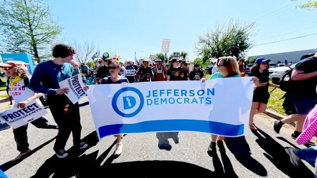 Group of people marching outdoors holding a large Jefferson Democrats banner and protest signs, some wearing colorful accessories, on a sunny day.